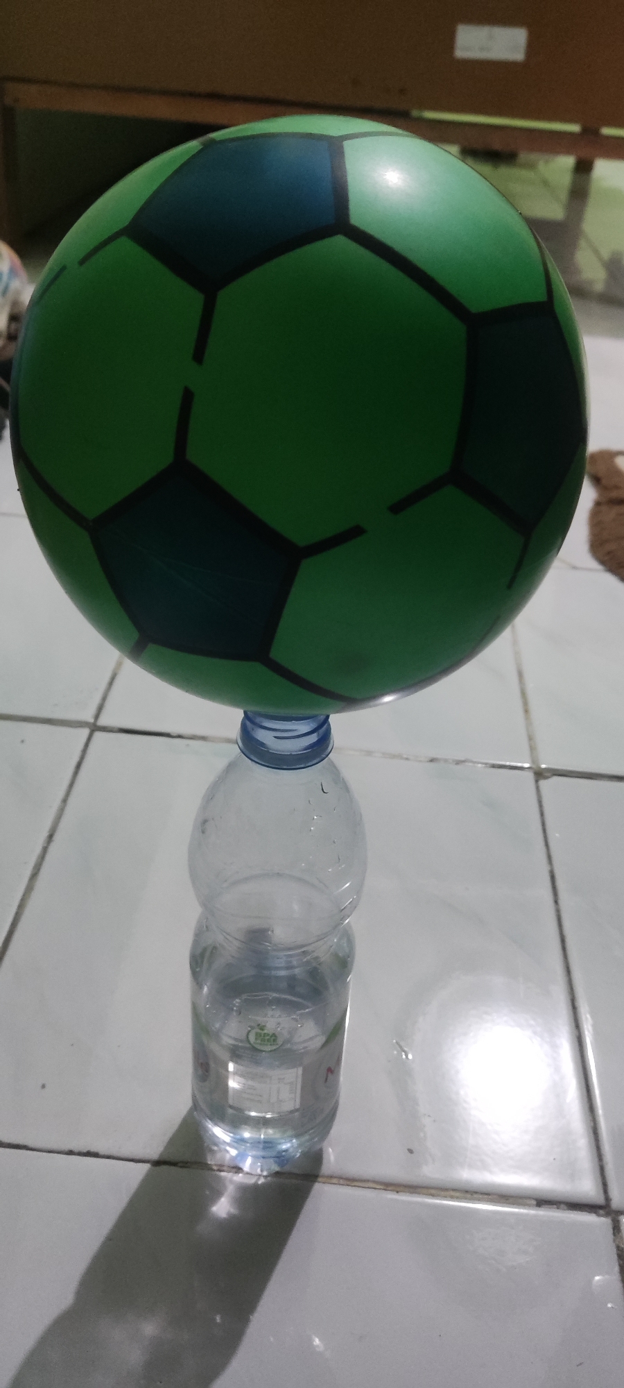 A green and dark blue soccer ball is balanced on the cap of a clear plastic water bottle. The ball has a classic hexagonal pattern. The bottle is partially filled with clear liquid. The scene appears to be indoors, on a tiled floor. The background shows a wooden surface, possibly a table or shelf, and some indistinct objects. The lighting suggests a dimly lit environment. The image was taken in Gebang, Indonesia.