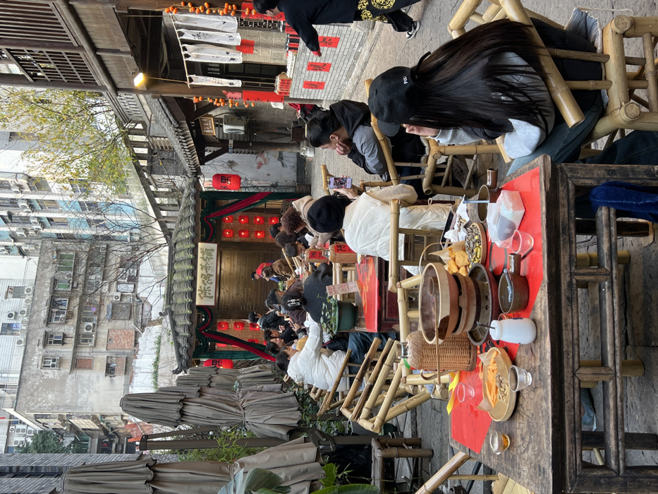 This image depicts an outdoor dining and tea area situated in an urban alleyway within Yuzhong District, China. The foreground features a rustic wooden table with a red tabletop, holding a multi-tiered clay hot pot or steamer over a burner. Surrounding the pot are various snacks including fried items, nuts, and dried fruit, alongside teacups, small bowls, a white pitcher, and a woven thermos. A person with long dark hair, wearing a black baseball cap and a grey hoodie, sits with their back to the viewer at a bamboo chair at this table, facing towards the scene.
Beyond this table, multiple other individuals are seated at similar wooden tables with bamboo chairs, engaged in eating, drinking, and social interaction. Many are observed looking at or using mobile phones. In the mid-ground, a person wearing a white vest is seen holding a smartphone, seemingly capturing the scene.
The environment is characterized by a blend of traditional Chinese architecture and modern urban elements. A prominent traditional-style building with a dark tiled roof and the sign "陈福茶铺" (Chen Fu Tea Shop) is visible in the center, adorned with red lanterns and festive red banners. To its left, several large, folded tan umbrellas are stored upright. To the right, another traditional wooden structure is decorated with white banners, red papers bearing Chinese characters, and strings of dried persimmons. In the background, modern, multi-story residential buildings with numerous air conditioning units rise above the traditional structures. The ground appears paved, and the lighting suggests an overcast or hazy daytime setting. A person in black attire with a patterned lower garment walks away to the right of the frame.
