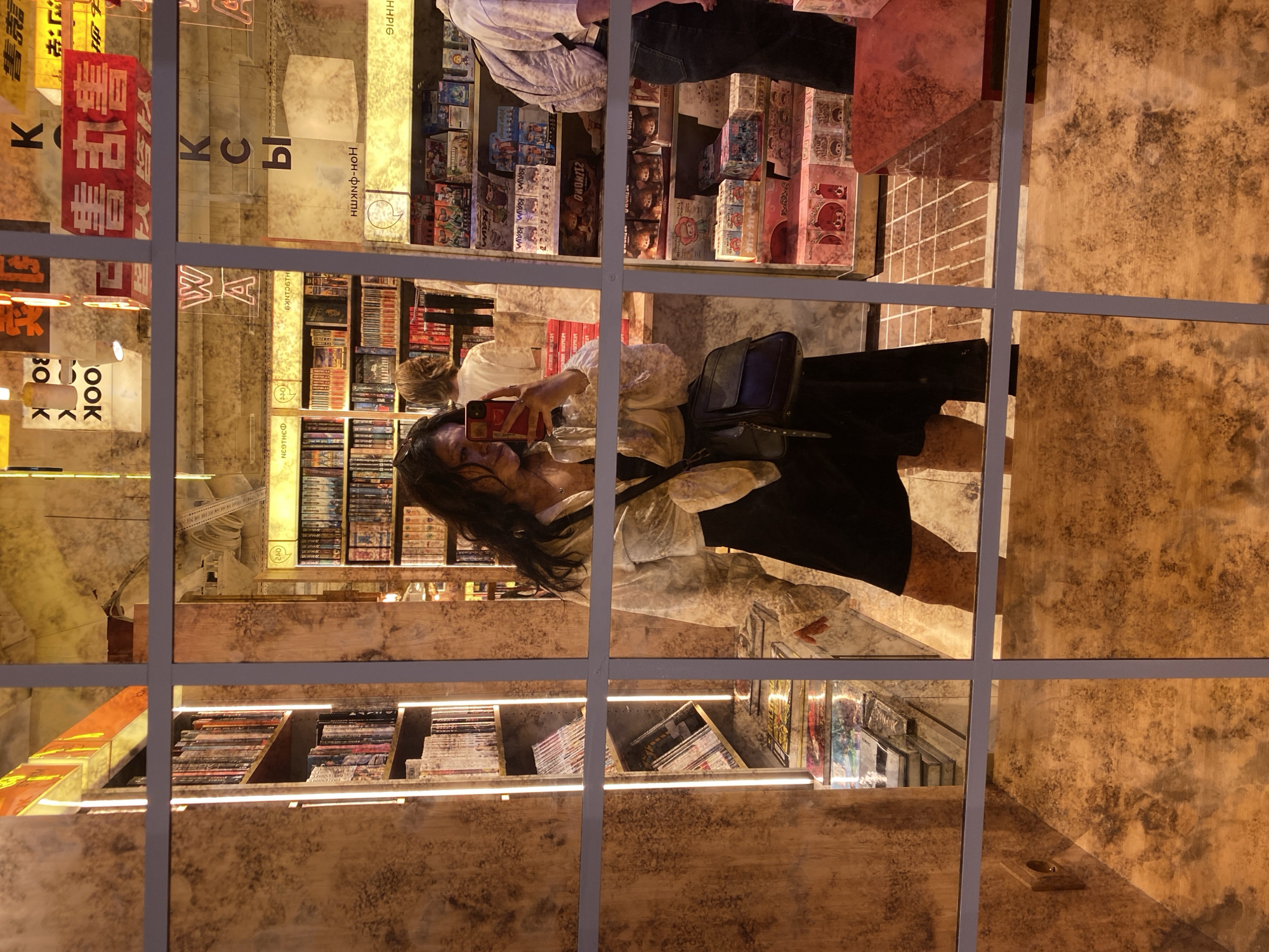 This is a mirror-image photograph taken inside a bookstore. A woman is taking a selfie reflected in a grid of mirrors. She is wearing a white patterned blouse and a black dress, and carrying a black purse. The background shows bookshelves filled with books, some of which appear to be manga or graphic novels. There are also display shelves with various merchandise, including boxes with colorful packaging. The overall aesthetic of the bookstore is warm and somewhat rustic, with wood-paneled walls. The reflection of street signs is visible in the upper part of the image, with Chinese characters and English letters, including "BOOK." The lighting appears to be artificial, with some shelves illuminated by vertical LED strips. The reflection of a red stool or table is visible in the lower right portion of the image. Based on the street signs visible in the reflection, the location is likely Saint Petersburg, Russia.