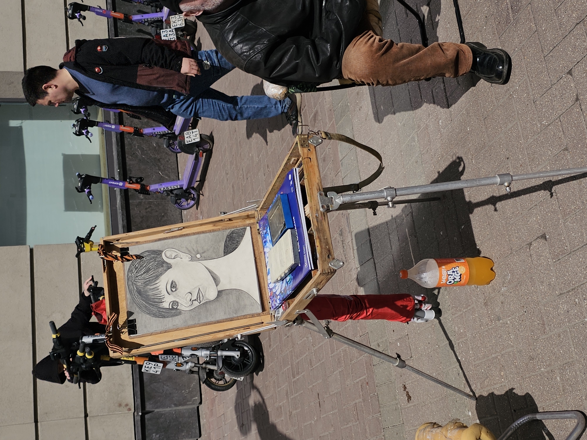 The image depicts an urban scene, likely in Moscow, Russia, as indicated by Cyrillic lettering on the license plates and the brand of the orange soda. The photo is taken at street level.
Foreground: An art easel is set up on the sidewalk. A portrait sketch of a woman is mounted on the easel. Below the easel, a small scooter-like motorized vehicle is parked. An open wooden box attached to the easel appears to contain art supplies. An orange soda bottle is lying on the ground.
Middle Ground: Several electric scooters branded with "ЮРЕНТ" (Yurent) are parked nearby. One person, dressed in jeans and a jacket, is walking past the scooters. A second individual, wearing brown trousers and black shoes, is visible in the frame.
Background: A building facade with windows and neutral coloring is visible behind the scooters and the artist's setup. The pavement is brick. The scene is set on a sunny day, with shadows cast by the objects and people.