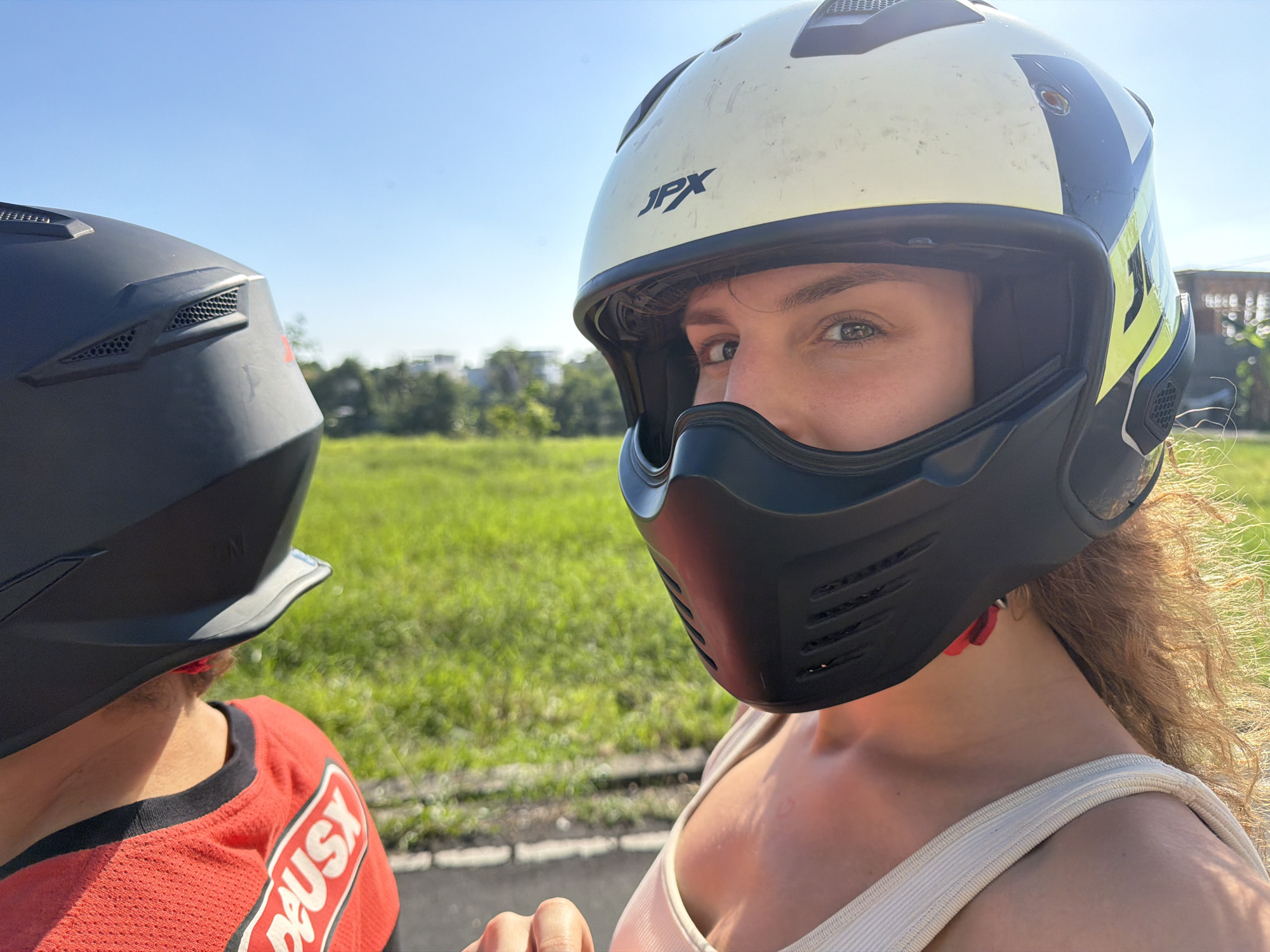 Two individuals wearing helmets are on a road in North Kuta, Indonesia, with a grassy field and trees in the background under a clear blue sky. The person in the foreground, wearing a white and black helmet with yellow accents and a light-colored tank top, looks towards the camera. The other person, wearing a black helmet and a red shirt with white lettering, is partially visible on the left side of the frame, looking away from the camera. The road appears to be paved, with a white line visible.