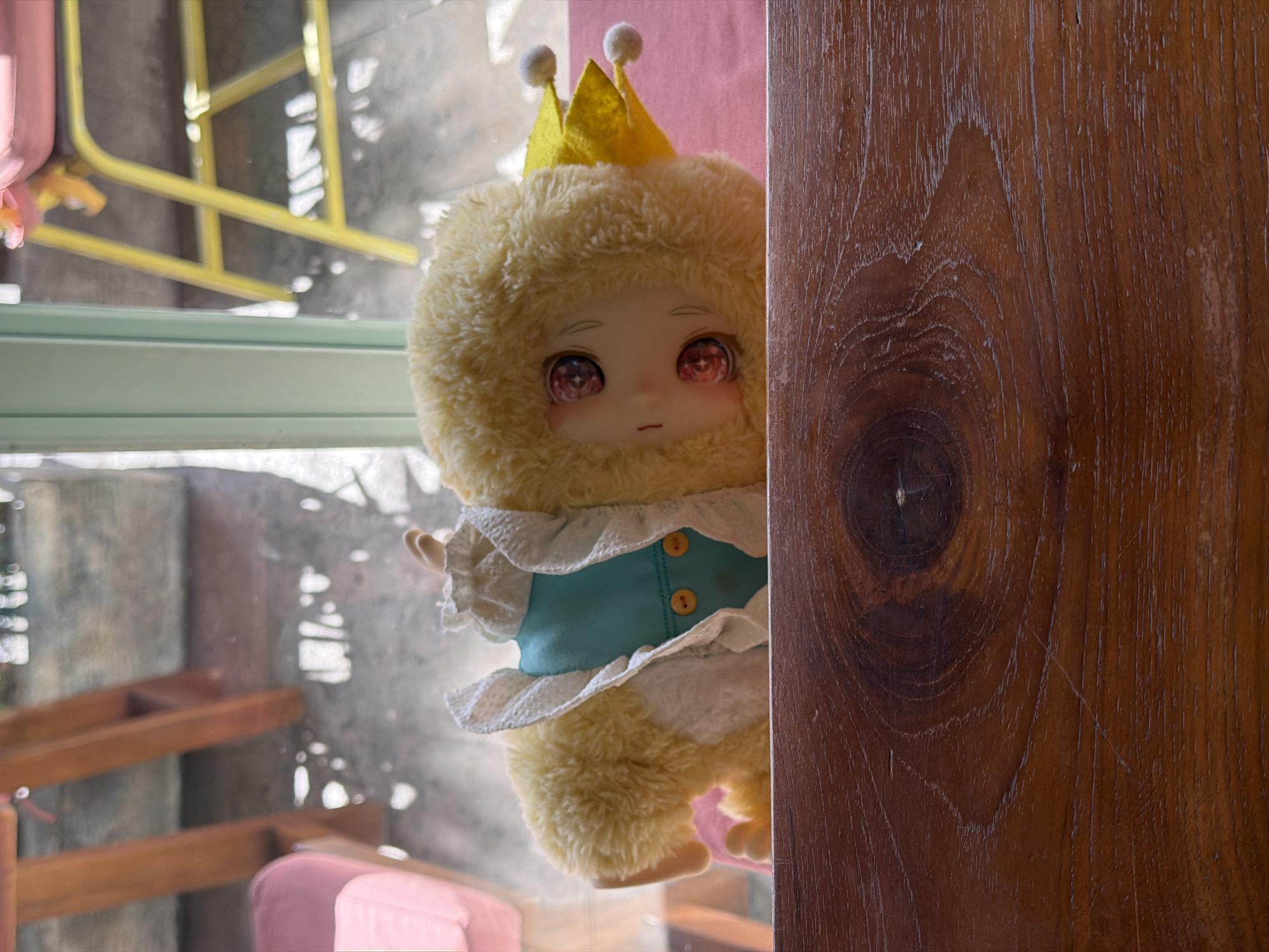 A fluffy, humanoid plush doll wearing a yellow crown and a blue and white ruffled garment peers from behind a dark wood surface. The doll has large, expressive red eyes. The doll's head and torso are visible, and it appears to be positioned near a window. Outside the window, a blurry scene includes a yellow metal structure, a pink object, and possibly chairs. The location is likely North Kuta, Indonesia. The scene is lit with soft, natural light.