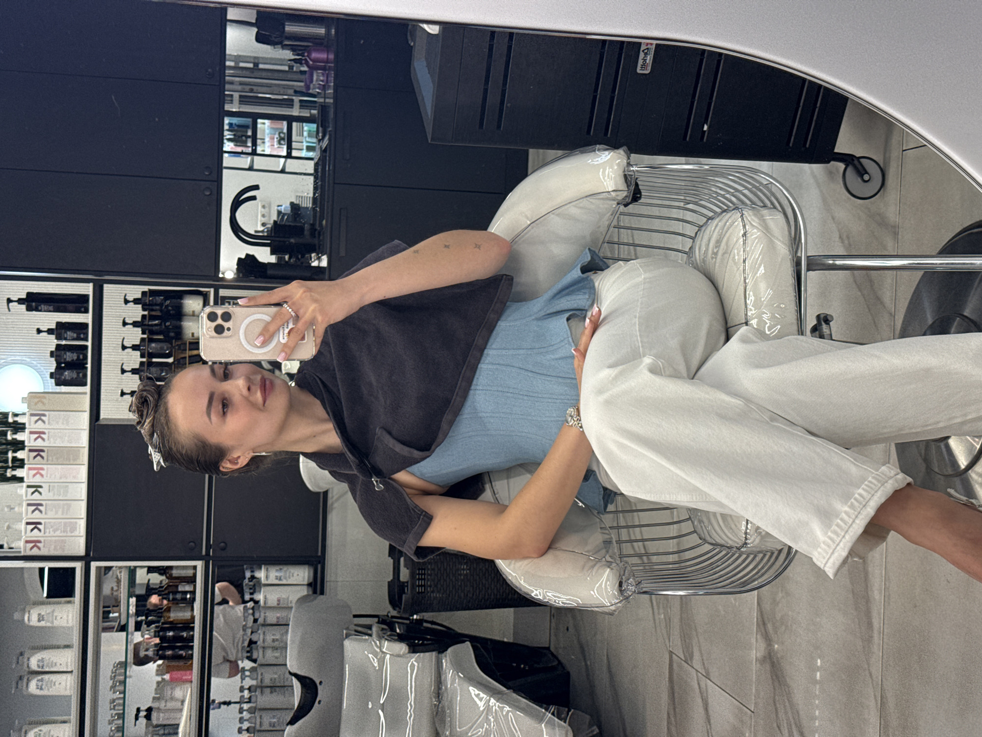 A woman is sitting in a chair in a salon and taking a selfie in a mirror. She is wearing a blue top and white pants and has a dark-colored towel around her shoulders. Her hair is up in a bun with a clip. She is holding a phone in her right hand, showing its back, likely to take a photo in the mirror. The salon has several shelves with hair products, a dark-colored cabinet, and styling chairs. The scene is likely located in Kyiv, Ukraine.