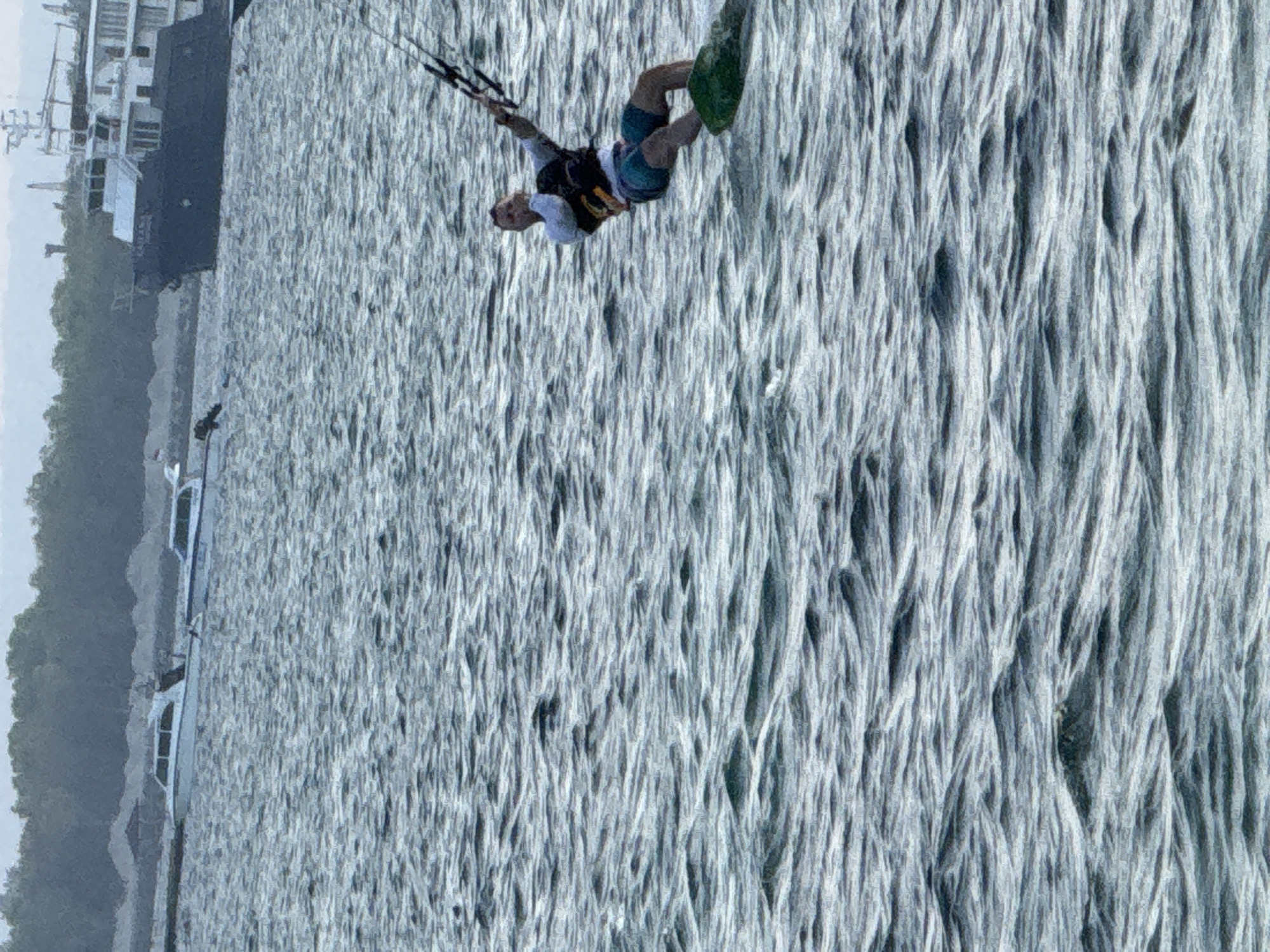 A person is kitesurfing on choppy water. The kitesurfer is wearing a white long-sleeved shirt, a dark vest, and blue shorts, and is holding onto a kite bar with both hands. They are leaning back, with their body angled and their right leg forward. The kite is visible in the upper right portion of the image. In the background, a large dark ship is docked near a tree-lined shore. Two smaller white boats are also visible in the water. The overall scene is set on a body of water with moderate waves. This image was likely taken in Denpasar, Indonesia, a city known for its coastal activities and proximity to the ocean.