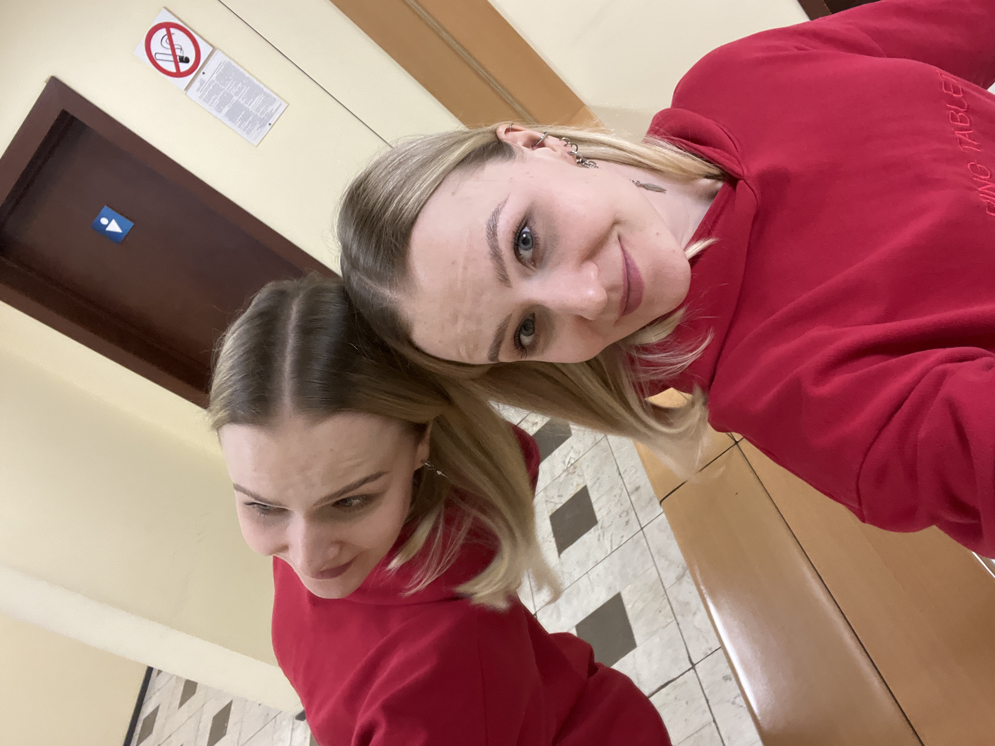 The image features two young women indoors, likely in a hallway or public area in Khimki, Russia. Both individuals are wearing matching casual red hoodies.
The woman positioned higher in the frame, with blonde hair and blue eyes, is looking directly at the camera with a gentle, friendly smile. Her left ear is adorned with several earrings, including hoops and a dangling charm. Partially visible text on her red hoodie reads "PING TABLE." Her head is tilted, resting lightly against the other woman's head.
The second woman, situated in the foreground, has lighter brown or darker blonde hair, possibly styled with small braids or sections, and is looking downwards and to her right, away from the camera. Her facial expression is not fully discernible.
The background consists of light cream-colored walls. On the left, a dark brown door features a blue and white restroom sign with male and female figures. Above the door, a prominent red and white "No Smoking" symbol is displayed next to a rectangular white sign containing small, illegible text. The flooring in the lower part of the image shows a checkered pattern of light and dark square tiles, adjacent to a smooth, light brown surface that might be a bench or part of the floor. The photograph is taken from an angled, slightly elevated perspective, suggesting a casual selfie or candid shot, with bright indoor lighting.