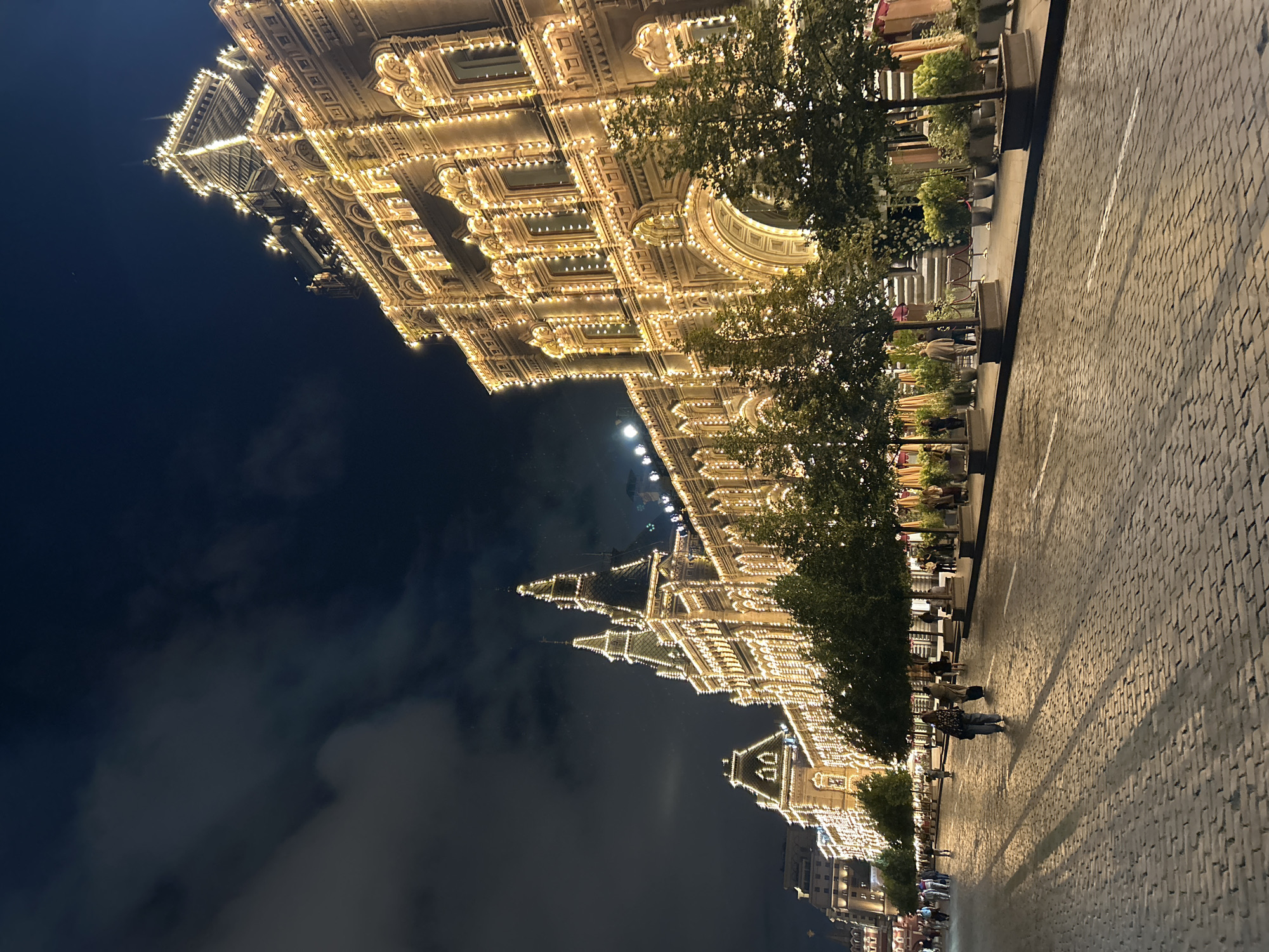 A night view of GUM department store on Red Square in Moscow, Russia, showcases its facade illuminated by numerous warm-toned lights. The cobblestone street in the foreground reflects the lights and the shadows of people walking on it. Several trees are planted along the sidewalk, adding a touch of greenery against the illuminated building and the dark night sky. A few people are visible on the street, walking and gathered near the entrance of the building. The architecture of GUM, with its intricate details and multiple towers, is highlighted by the festive lighting. The sky is dark with some clouds visible.