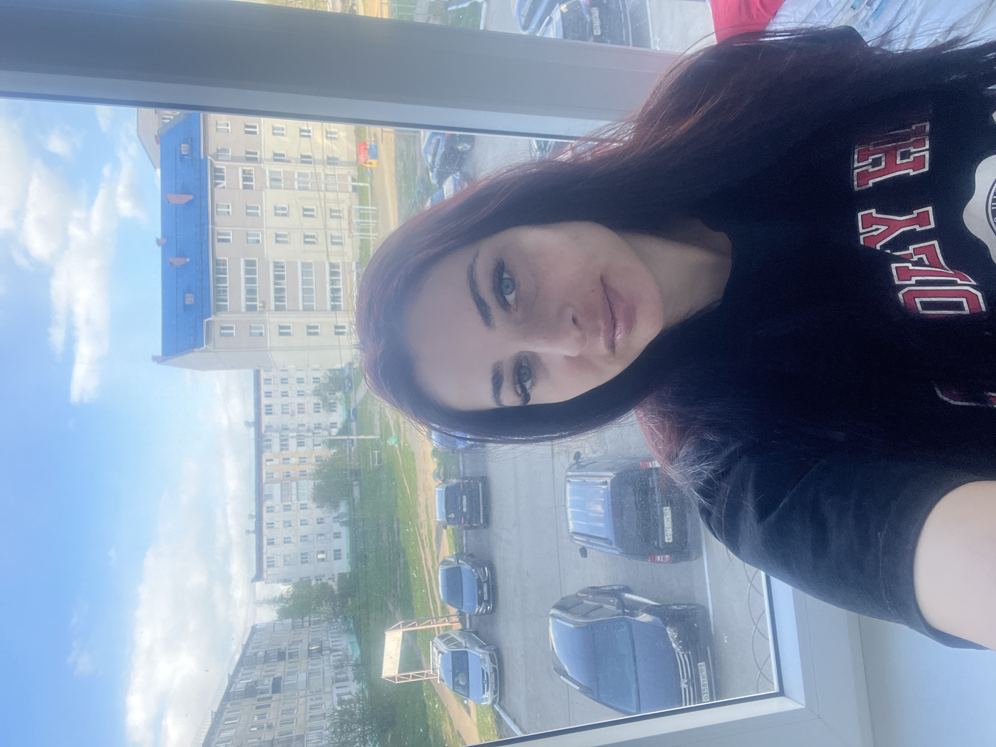 A woman is taking a selfie indoors, presumably near a window, with a cityscape visible outside. She has dark hair, light eyes, and is wearing a dark-colored shirt.
The background shows a street with parked cars and several apartment buildings. The architecture suggests an urban residential area in Chaikovsky, Russia. The sky is partly cloudy. A basketball hoop is visible in the courtyard.