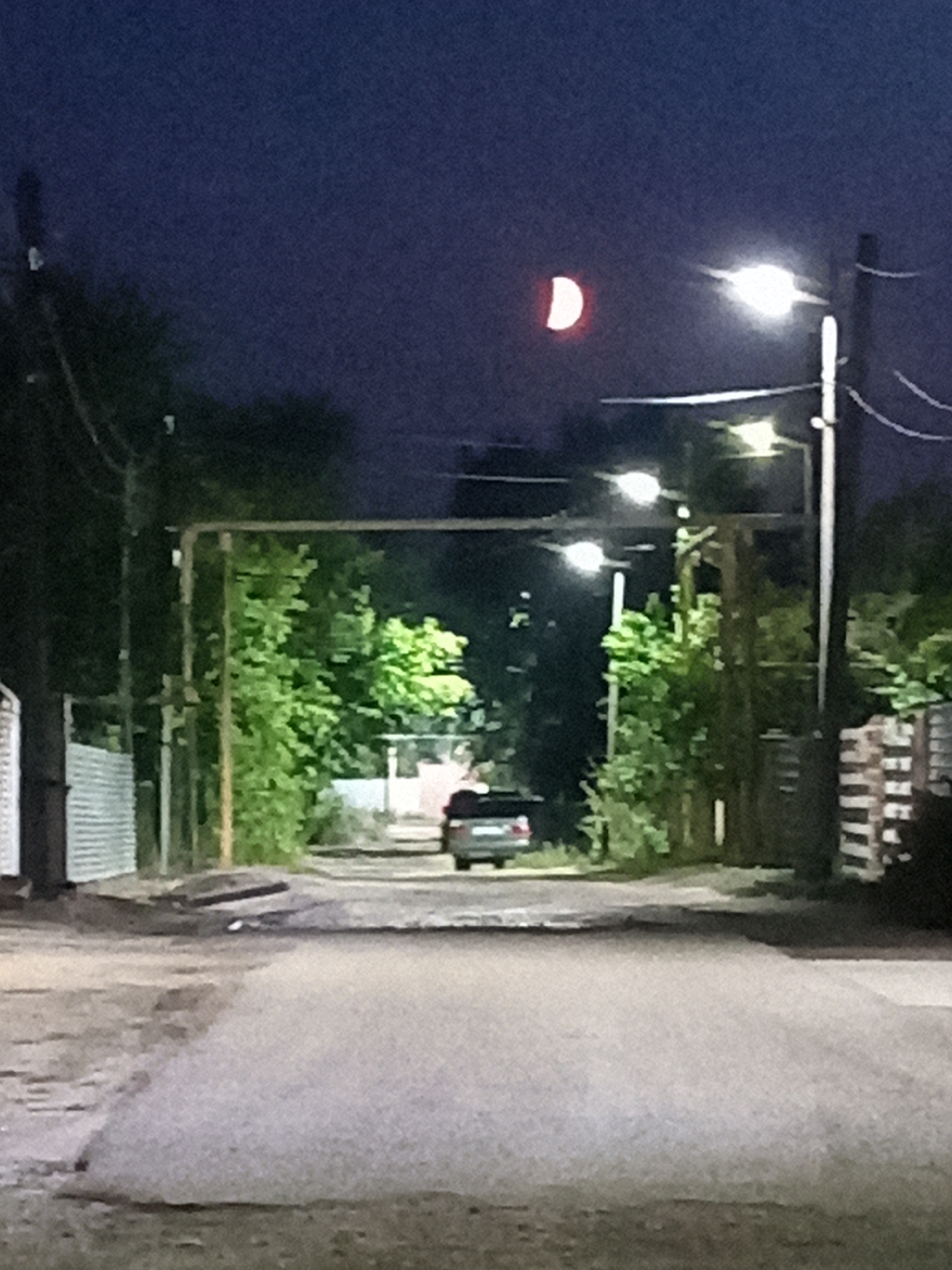 A crescent moon is visible in the dark blue night sky above a street in Rudny, Kazakhstan. The moon is lit by the sun from the right and appears as a bright, red-orange sliver. Streetlights illuminate the scene, casting a glow on the asphalt road and surrounding trees. A silver car is parked on the right side of the road, and a white fence runs along the left side. The overall atmosphere is quiet and peaceful, with the natural beauty of the night sky complementing the urban landscape.