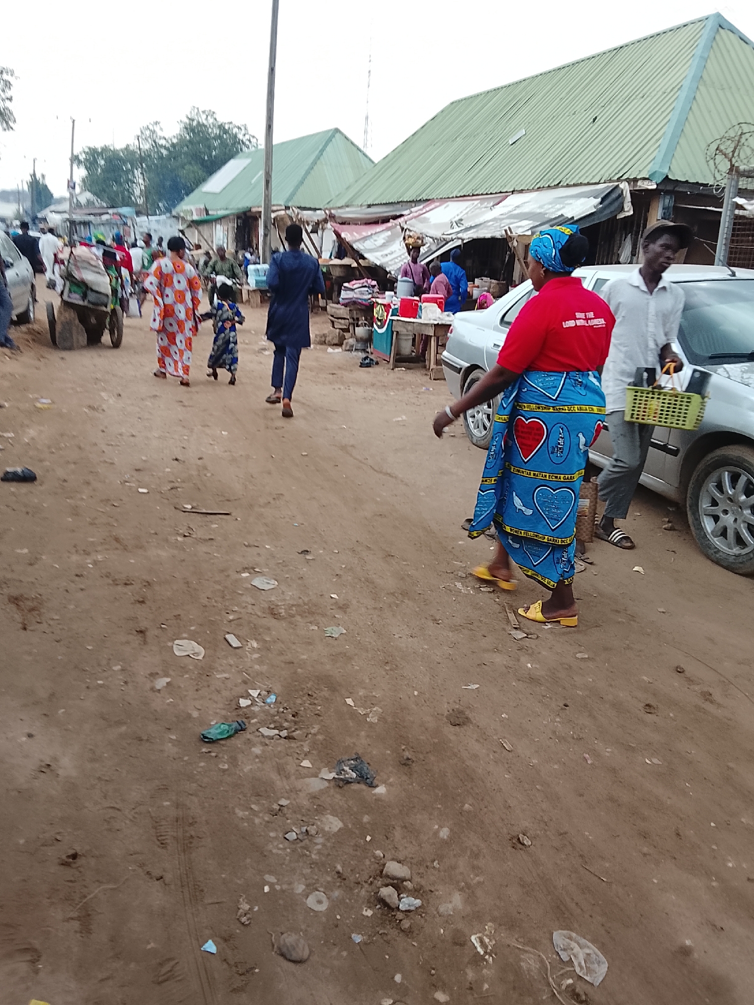 The image shows a dirt road in what appears to be a market area. Several people are walking along the road. A woman in a red shirt and blue patterned skirt is walking towards the right side of the frame, while a man in a white shirt and sandals is walking beside her, carrying a green basket. Another group of people, including a woman in an orange and white patterned dress and a child, are walking further down the road away from the camera. There is a car parked on the right side of the road. Stalls with goods are set up along the road. The buildings have green roofs. There is litter scattered on the ground. The location is likely a market in the city of Unknown, Unknown.