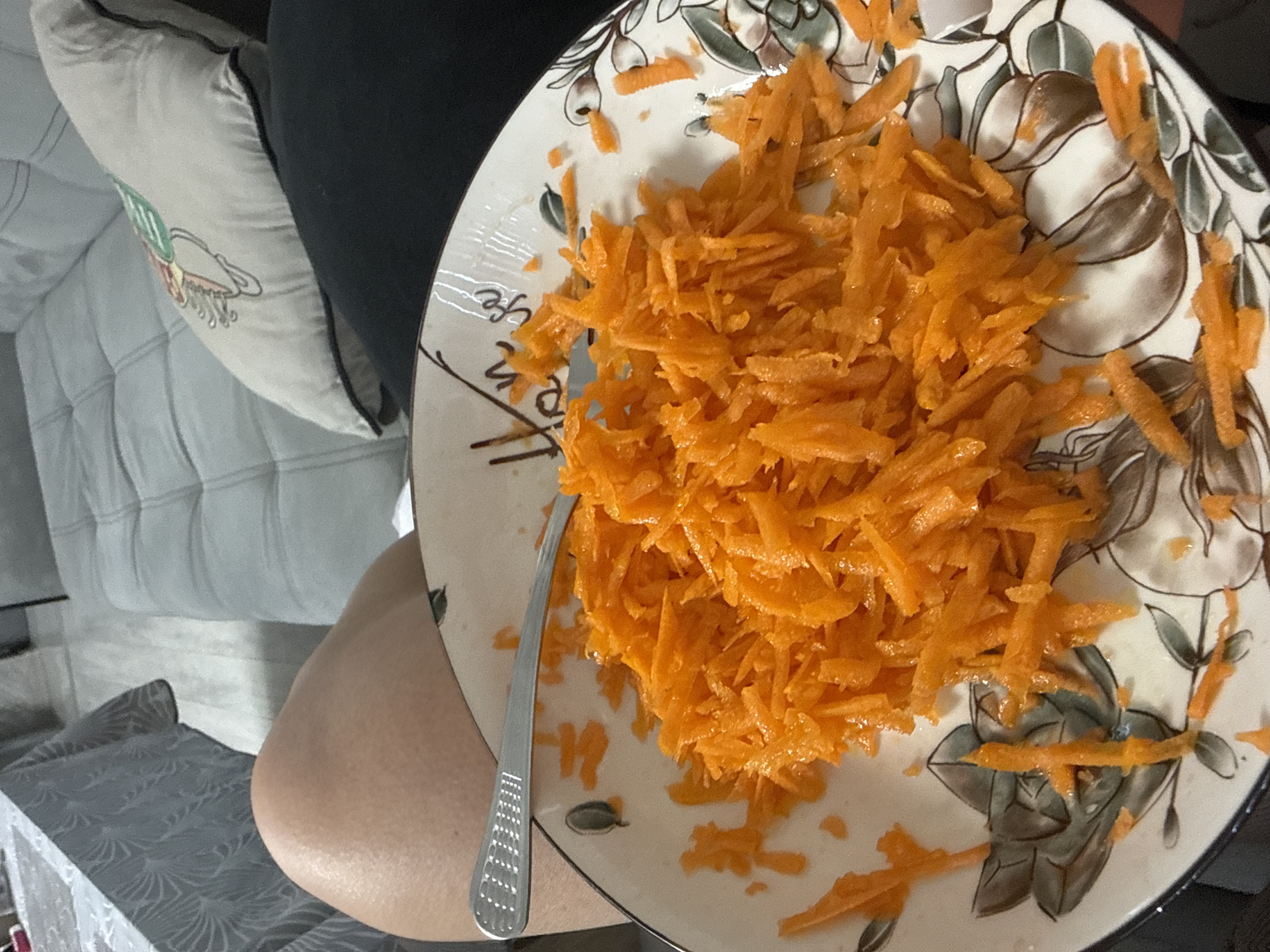 A close-up shot captures a plate filled with shredded carrots, accompanied by a fork resting on the edge. The plate itself is adorned with a decorative pattern featuring floral motifs in shades of grey and brown. In the background, a portion of a person's leg is visible, suggesting they are seated. Further in the background, a grey tufted sofa and a grey tablecloth on a table are discernible, hinting at an indoor setting, likely a home. The scene appears to be within the city of Rogosh, Bulgaria, though no specific landmarks confirm this location. The overall impression is one of a casual meal or snack being prepared or consumed.