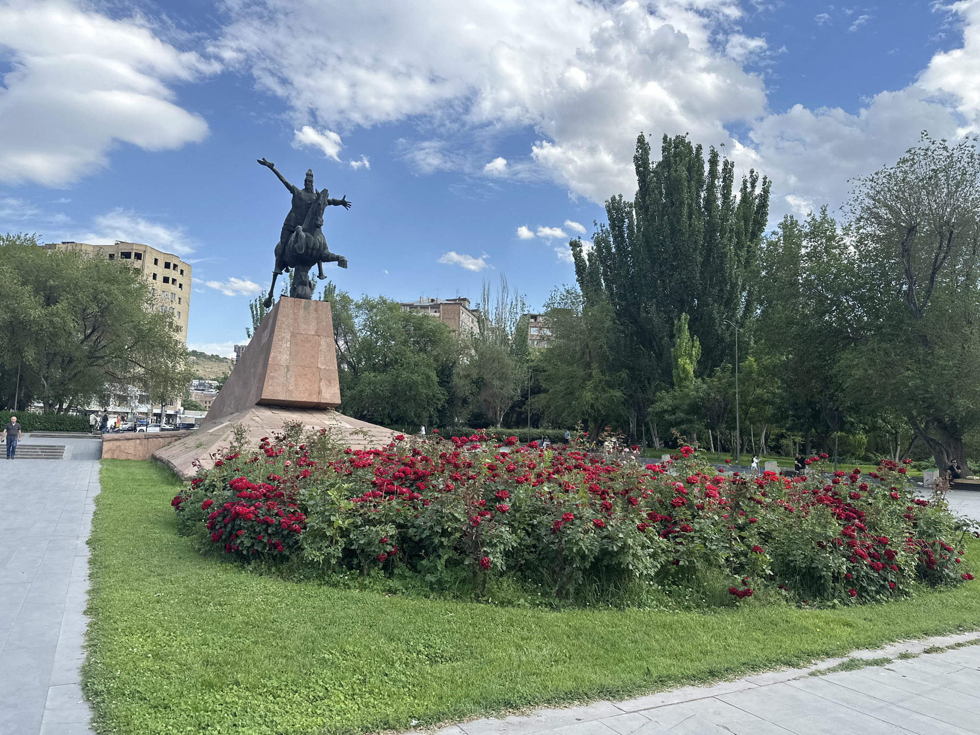 A bronze equestrian statue, depicting a figure with outstretched arms on a rearing horse, stands prominently on a tall, angled reddish-brown stone pedestal. The statue is the focal point of a public park or square, likely located in Yerevan, Armenia, as per the provided context.
Surrounding the monument is a lush, well-maintained lawn of green grass. In front of the pedestal, a large, crescent-shaped flowerbed bursts with vibrant red roses in full bloom, adding a striking color contrast to the scene. Various trees with dense green foliage, some tall and slender, others with broader canopies, frame the park, particularly on the right side and in the background.
The sky is bright blue with scattered white cumulus clouds, indicating a clear, sunny daytime. In the distance on the left, an old, multi-story building with numerous missing windows is visible, suggesting urban surroundings, along with other buildings further back. Paved walkways are visible on the left side of the park, where a lone man is seen walking away from the camera. Other indistinct figures are sparsely distributed in the background among the trees and buildings. The overall atmosphere is calm and pleasant, showcasing a common urban green space. No discernible text is visible in the image.