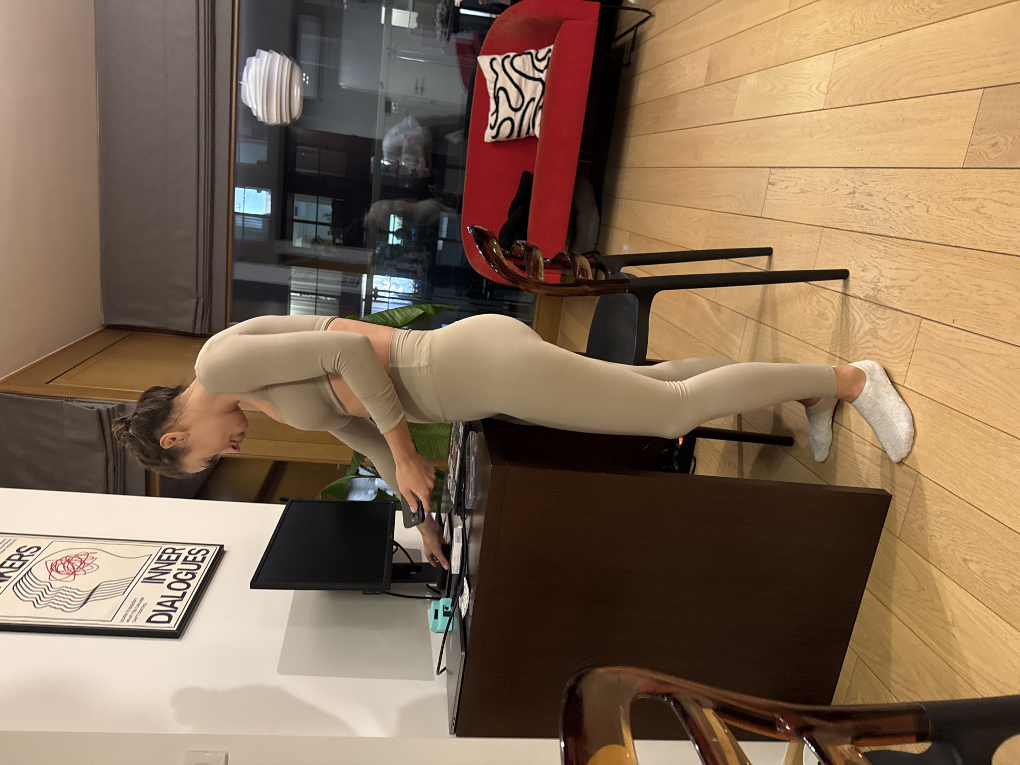 A woman in a light beige athletic outfit is performing a modified plank with her forearms resting on the edge of a dark brown desk. She is facing towards the desk and looking down at it. She is wearing white socks. The desk has a black computer monitor on it, various wires and electronic accessories, and a potted plant. On the wall is a framed poster.
The location appears to be an apartment or home office. A red chair with a decorative pillow is visible in the background, as is a window with a view suggesting an urban setting. The floor is made of light brown wood. The scene is likely in Warsaw, Poland, based on the location and surrounding environment.