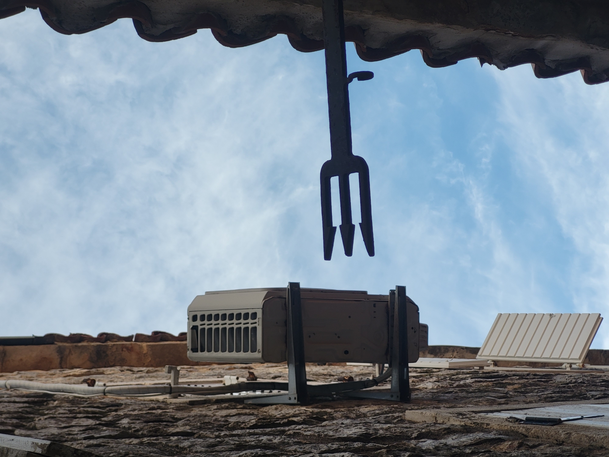 The image captures an upward view of a building's exterior against a bright, partly cloudy blue sky. A prominent, dark metal, three-pronged fork-like object hangs from an overhanging tiled roof in the upper center, with a small curved hook detail on its shaft. Directly below this object, an outdoor air conditioning unit or similar electrical appliance is mounted on a bracket against a textured, weathered wall. This unit is light-colored with visible vents on its left side.
In the upper part of the frame, the underside of a tiled roof edge is visible, made of reddish-brown clay tiles. Below the mounted unit, a lower roof or wall surface is visible, covered in what appears to be old, rough, dark-toned tiles or stone. White piping or cabling runs horizontally across this lower surface. Further to the right on this lower level, another white, louvered rectangular unit is present, angled upwards, possibly an exhaust vent. The overall scene suggests an old or traditional building. The lighting indicates it is daytime. No people or text are visible. The setting is consistent with a Mediterranean town like Budva, Montenegro, known for its old architecture and tiled roofs.