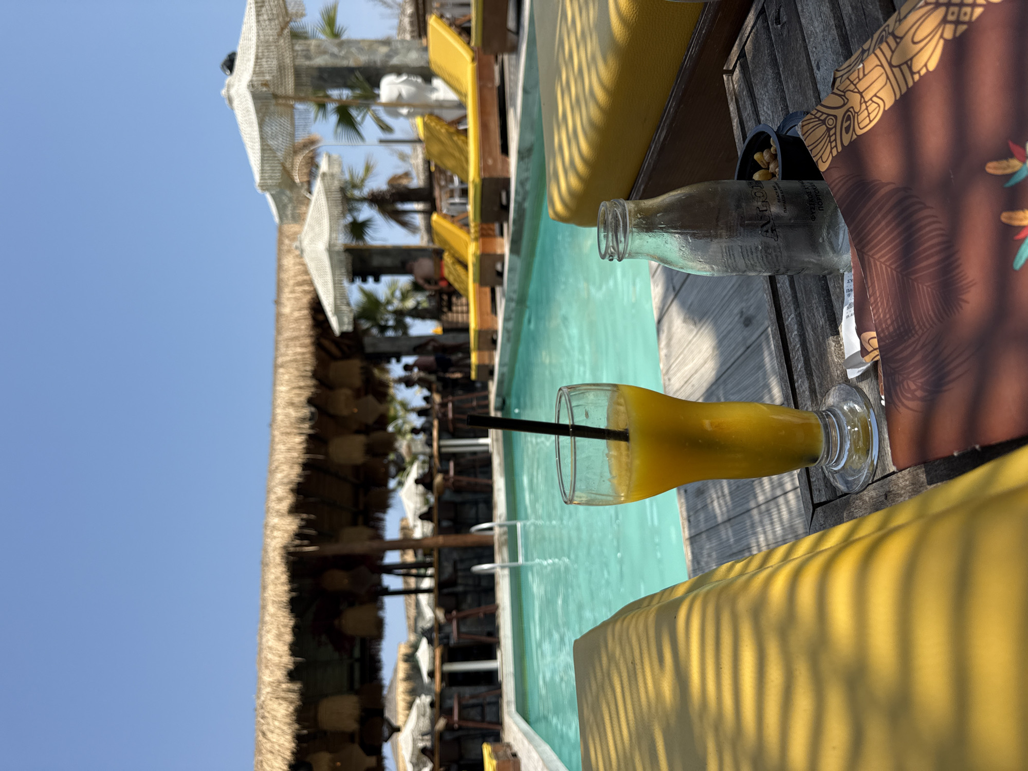 This is a medium shot of a bright, sunny day at a beachside resort. In the foreground, a wooden table holds a clear glass filled with a yellow beverage and a black straw, a clear glass bottle, and a small black bowl containing nuts. The table is positioned in front of a yellow lounge chair, casting dappled shadows on its surface. To the left, a swimming pool with turquoise water is visible, with a ladder submerged in it. Beyond the pool, a bar area with thatched roofing and hanging lights is set up, with several people seen in the background. Yellow lounge chairs are also scattered around the pool area, some with white umbrellas for shade. In the far background, palm trees and more umbrellas can be seen, suggesting a tropical or Mediterranean setting. The sky is a clear, bright blue. The scene depicts a relaxing atmosphere at a vacation spot.