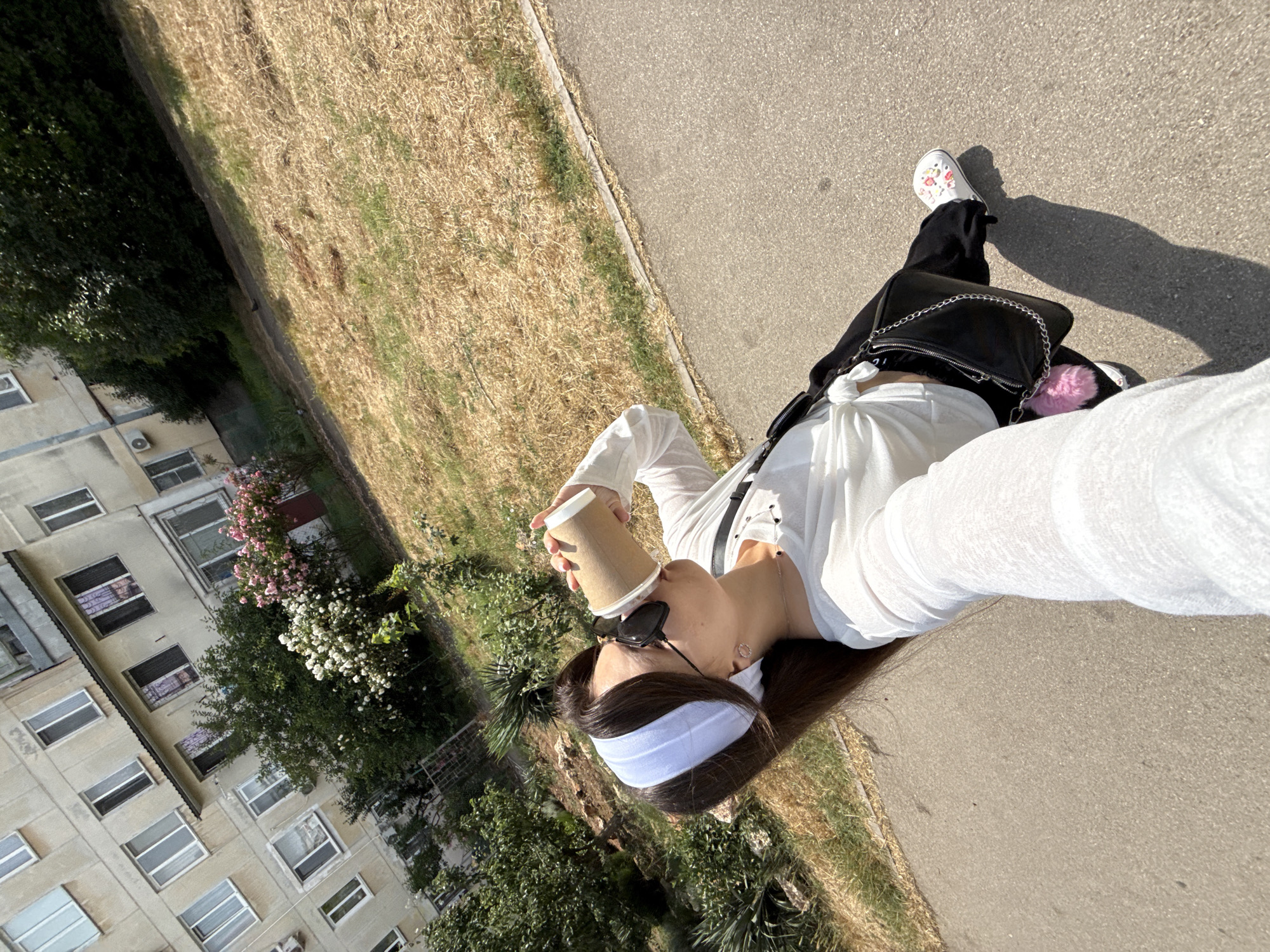 A young woman is standing on an asphalt path, holding a disposable coffee cup to her lips, and taking a selfie with her left hand. She has long dark hair pulled back with a white headband and is wearing sunglasses. She is dressed in a white long-sleeved shirt tied at the waist, black pants, and white shoes with colorful embellishments. A black purse with a silver chain strap and a pink pom-pom hangs from her left shoulder. The background shows a multi-story apartment building with many windows, lush green trees, and a patch of dry, brown grass. The scene appears to be outdoors on a sunny day.