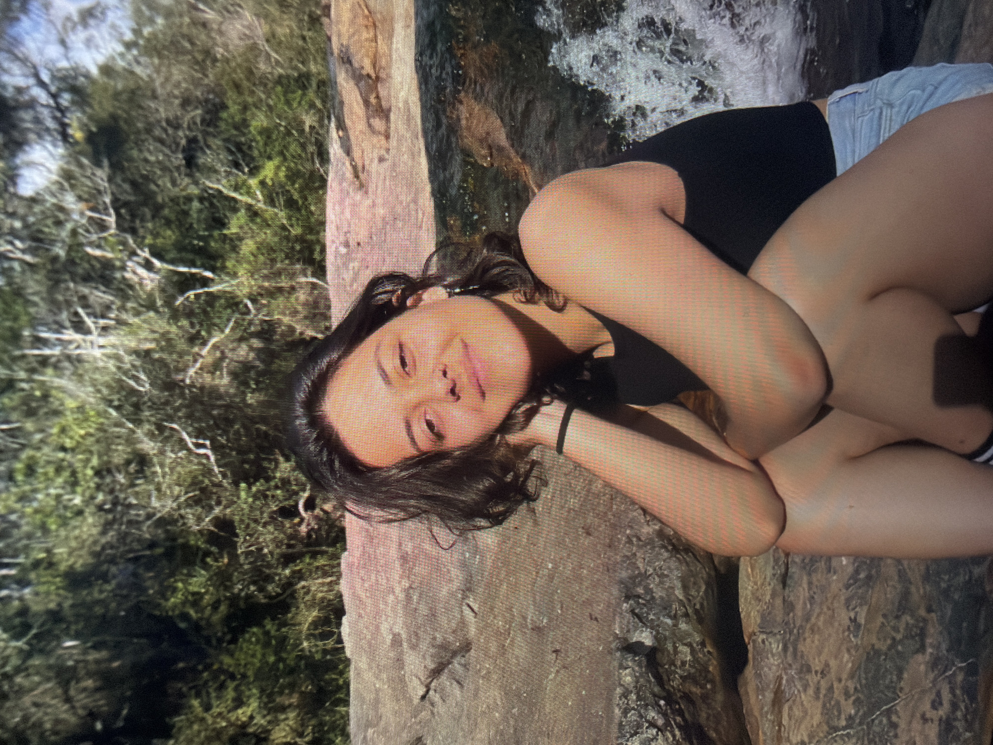 A young female, appearing to be in her late teens or early twenties, is seated outdoors, facing the camera with a slight smile. Her dark, wavy hair is parted to the side, framing her face. She wears a black tank top, light-colored denim shorts, and a black elastic band on her right wrist. Her right elbow rests on her right knee, with her right hand positioned near her chin. Her left arm is bent across her lap, with her left hand resting on her left knee. She is positioned on a large, textured rock formation, which appears to be part of a streambed or rocky bank. To her right, water cascades over rocks, forming a small waterfall or rapids. The background consists of dense green foliage and trees, indicating a natural, possibly wooded or forested environment. Natural sunlight illuminates the scene, casting shadows on her face and body, suggesting a bright day. The image exhibits visible pixelation and a grid pattern, characteristic of a photograph taken of a digital screen.