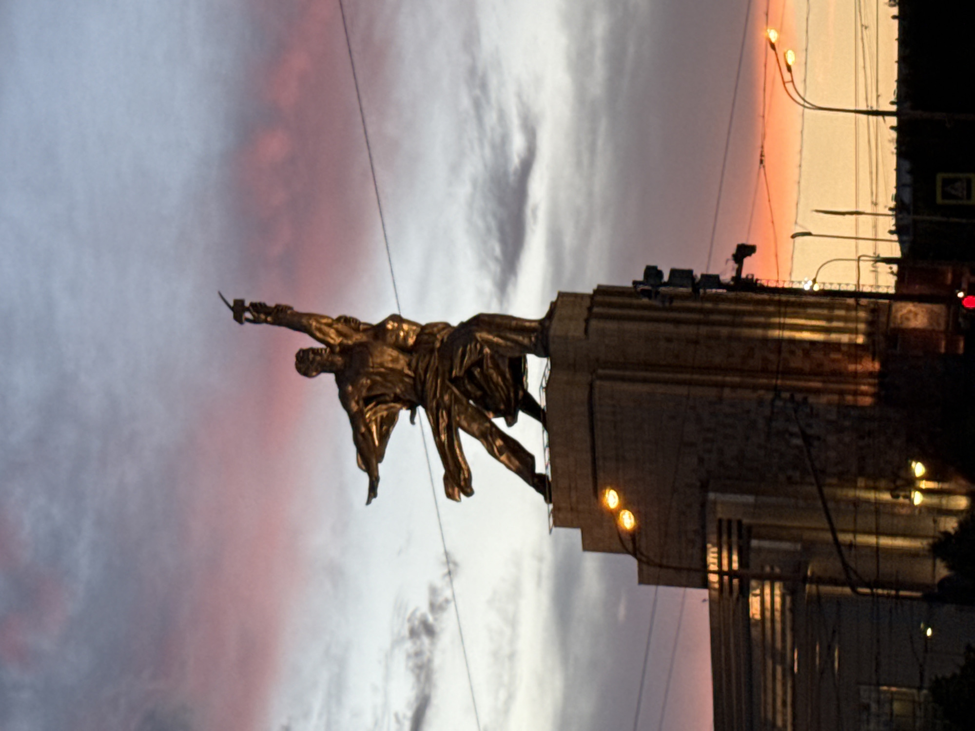 This is a nighttime, rotated image of a large bronze sculpture of two figures, likely a male and a female, with the male figure holding a hammer and the female figure holding a sickle. They are depicted in a dynamic, upward-reaching pose, symbolizing labor and progress. The sculpture is situated in Moscow, Russia, and is a prominent landmark.
The sculpture stands atop a substantial, multi-tiered pedestal or building, partially visible on the right side of the frame. The background consists of a dramatic sky, illuminated by a sunset or twilight glow, with soft hues of pink, orange, and grey clouds. The lighting suggests it is either early morning or late evening. Several streetlights and electrical wires are visible, indicating an urban environment. The overall mood is one of grandeur and monumentality, capturing a sense of historical or ideological significance often associated with Soviet-era art. There is a traffic sign with Cyrillic text visible on the building.