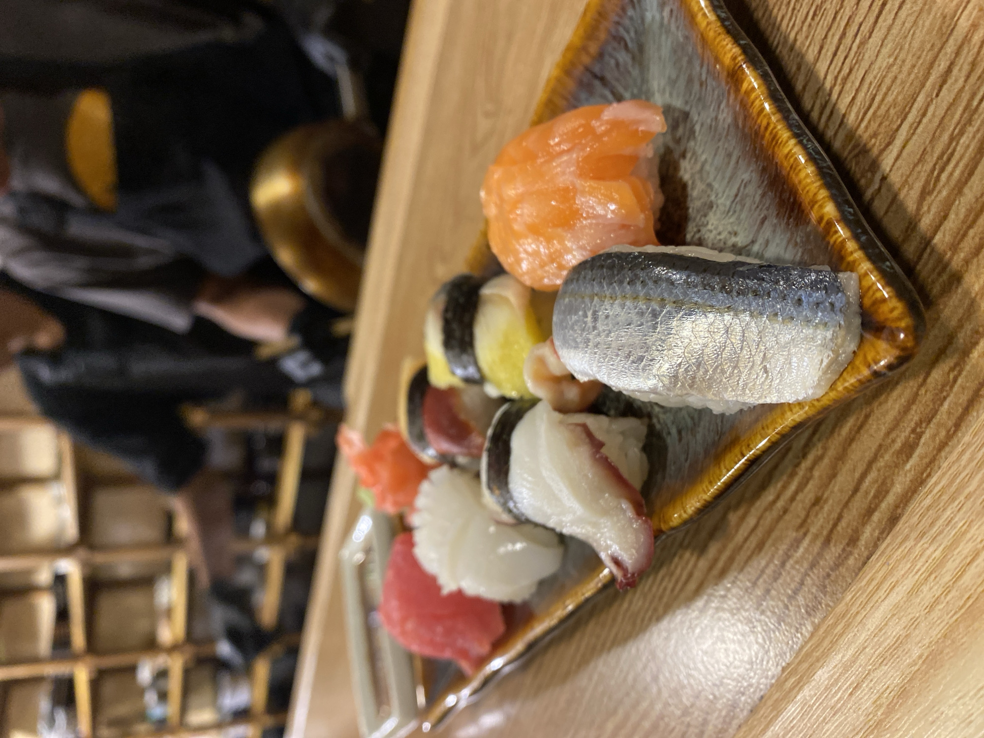 The media file displays a close-up of a plate of assorted sushi, set against a wooden surface. The sushi includes pieces with salmon, mackerel, octopus, and other unidentifiable seafood. The plate itself is rectangular with a dark, textured rim featuring a gradient of brown and black. In the background, a blurred image shows people and what appears to be shelving units, suggesting an indoor setting possibly a restaurant or home. One person's arm is visible, wearing dark clothing. The lighting suggests daytime, and the overall atmosphere is casual, focused on the food presentation. There is no visible text. The context of Da Lat, Vietnam, might imply a local interpretation or offering of sushi.