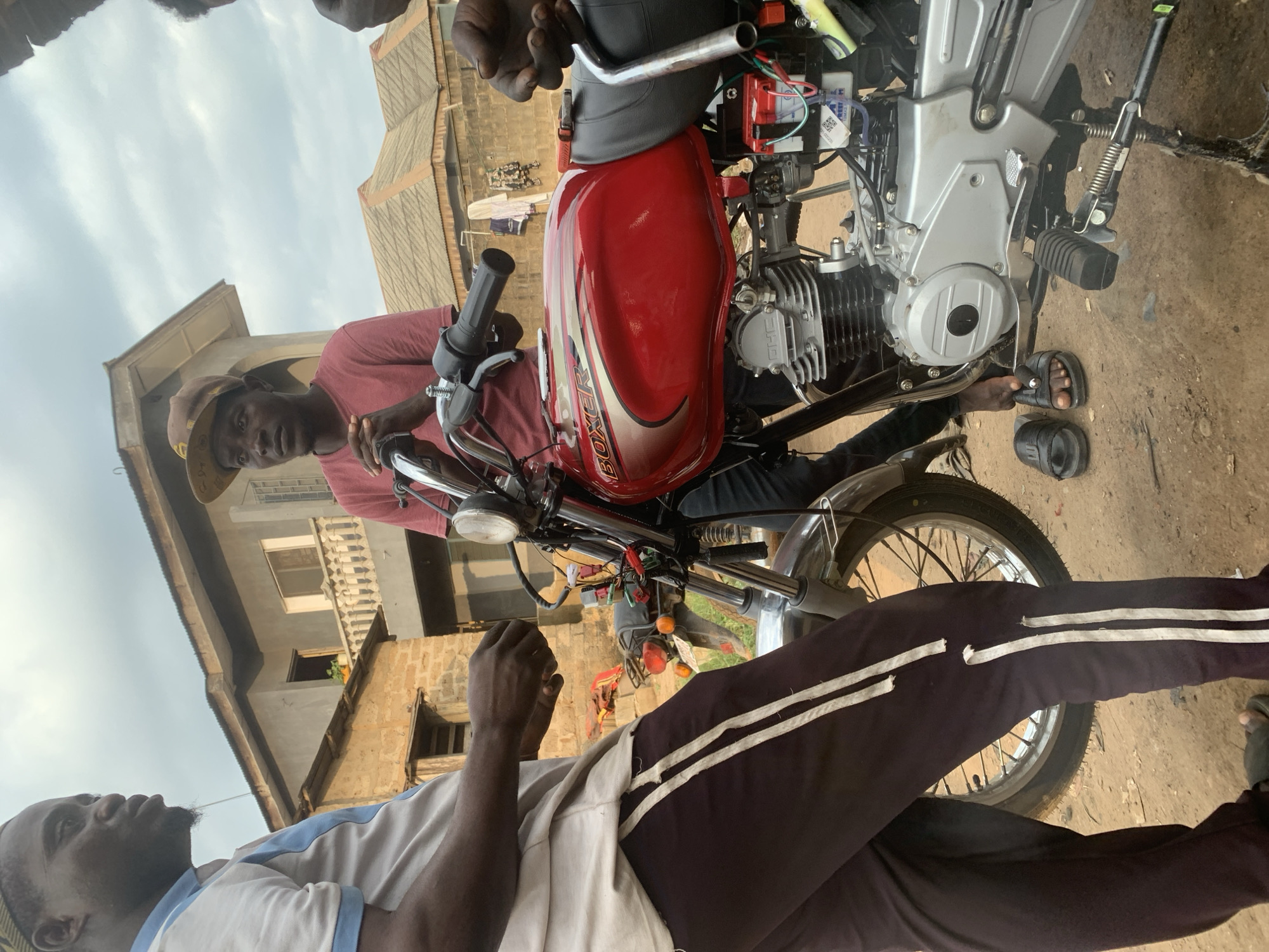 The image depicts a street scene in Shagamu, Nigeria, where two men are gathered around a red Boxer motorcycle. The man on the left, wearing a white and blue shirt and dark pants with white stripes, is in the foreground, his hands engaged near the motorcycle's fuel tank. To the right of him, a second man, wearing a red t-shirt and a brown baseball cap, stands next to the motorcycle, his hands resting on the handlebars. The motorcycle, a prominent subject, features a red fuel tank with "BOXER" branding. Electrical wires and a battery are visible on the side of the engine. The background reveals typical urban Nigerian architecture, including multi-story buildings with balconies and corrugated iron roofs, along with some laundry hanging outside. The ground is a mix of dirt and sand. The overall impression is of a casual interaction or a moment of work being done on the motorcycle in a residential area.