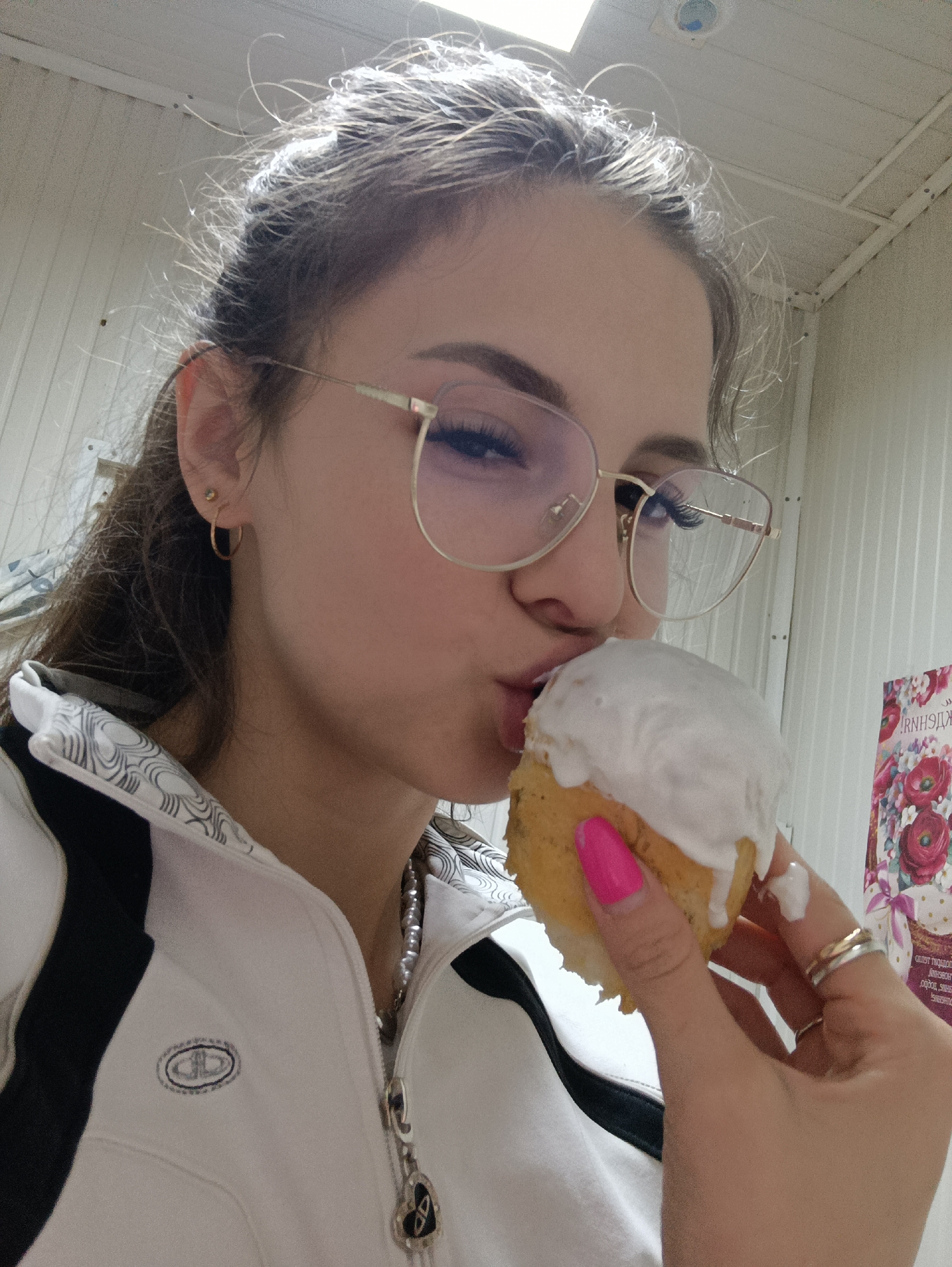 A young woman with brown hair, wearing gold-framed glasses and a white tracksuit with black stripes, is holding a small cake with white frosting. Her nails are painted bright pink, and she is wearing several rings. She appears to be taking a bite of the cake. The background consists of what seems to be the corner of a room or a hallway. There is a poster with flowers visible in the background.
The location is Прикамский, Russia.