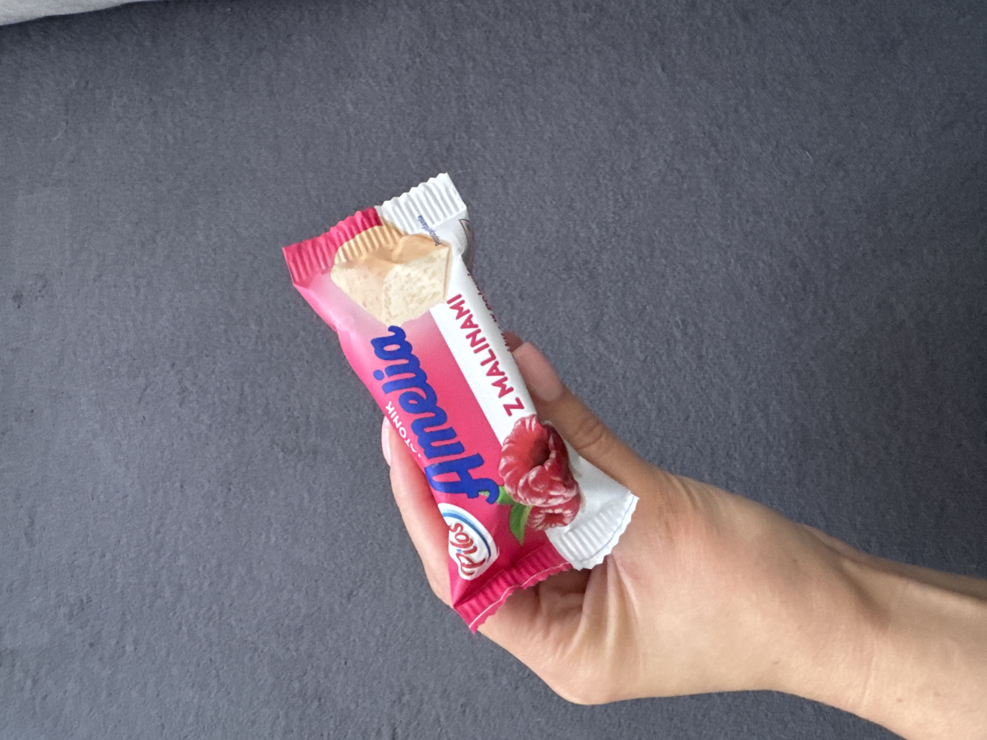 A hand is holding a wrapped candy bar with a pink and white wrapper. The wrapper has text that appears to be in Polish and images of raspberries. The background is a dark, textured surface. The image was taken in Zielona Góra, Poland.