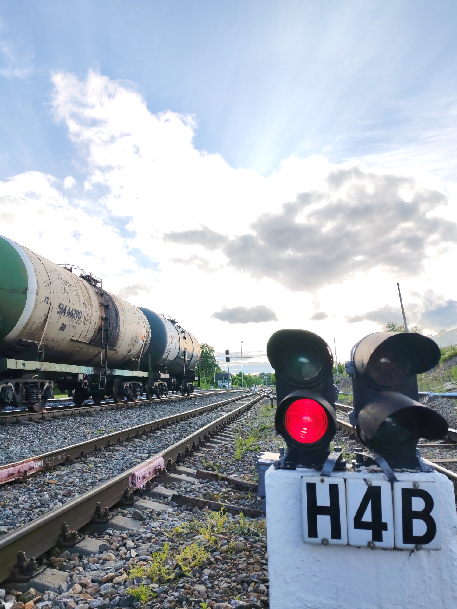 The image shows a railway scene in Bol'shie Mkhi, Russia. A train composed of tank cars occupies one set of tracks. A red railway signal, labeled "H4B," is prominently displayed in the foreground, indicating a stop condition. The tracks are embedded in gravel. The sky is partly cloudy.