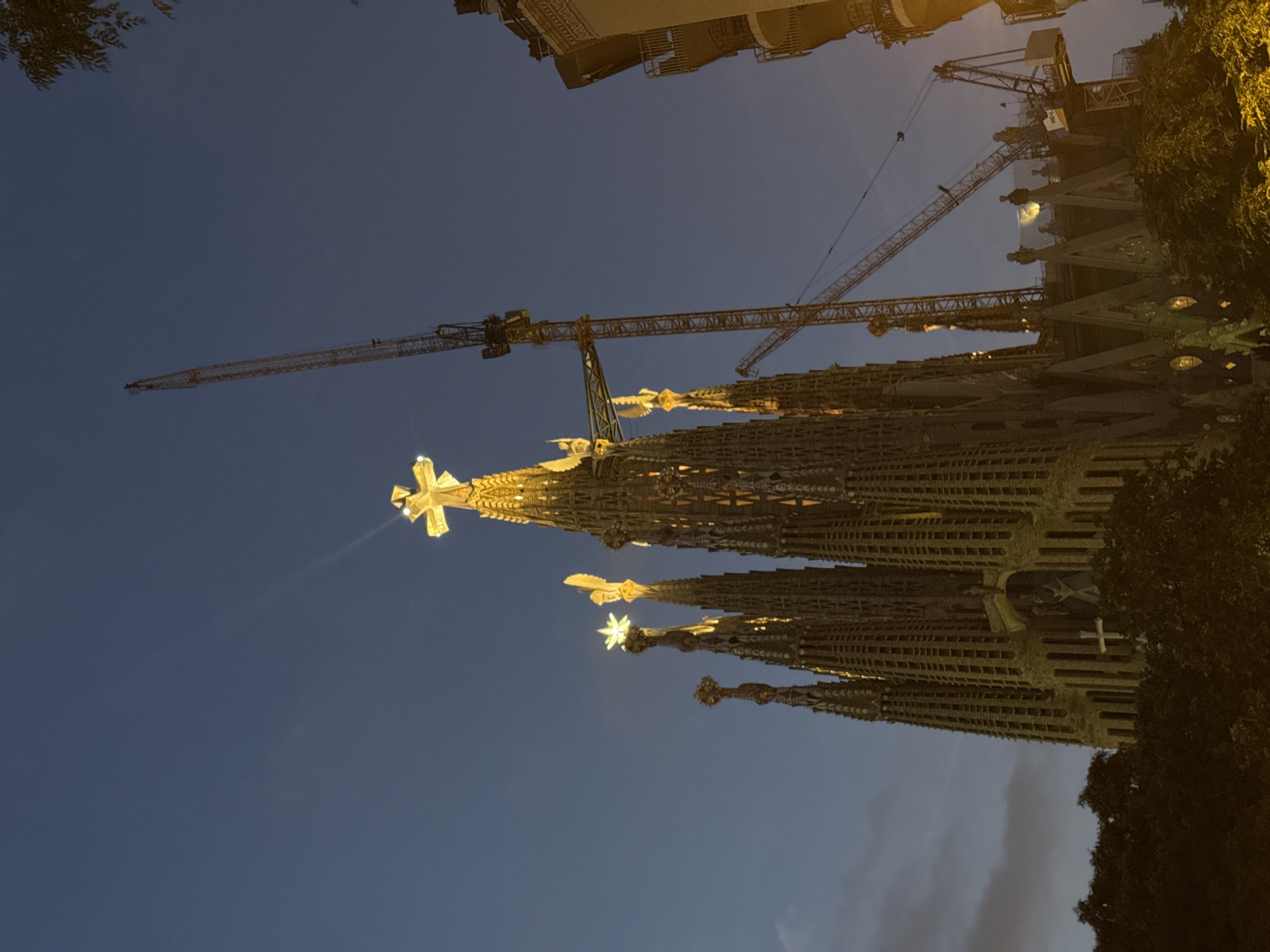The image captures a low-angle, night-time view of the Sagrada Familia basilica in Barcelona, Spain, under construction. The basilica's spires, illuminated by warm yellow lights, dominate the center and right of the frame. Several spires are topped with illuminated crosses and a star. Two large construction cranes are visible against the dark blue sky, one extending high above the basilica, and another, smaller one, positioned lower down. The detailed stonework of the basilica's spires is visible, with a faint luminescence emanating from within some sections. Trees with dark foliage partially obscure the base of the structure in the foreground. To the far right, a portion of a modern building with balconies is visible. The sky is a deep, clear blue, transitioning to a lighter shade towards the horizon. The overall impression is of an ongoing architectural project under artificial light at dusk or night.