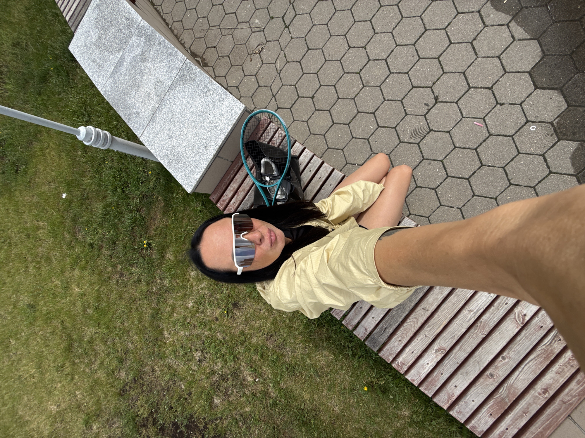 A woman is sitting on a wooden bench on a sunny day in Chaikovsky, Russia. She has long dark hair and is wearing large white sunglasses and a light yellow jacket. A teal tennis racket is lying on the bench next to a black bag. The bench is located next to a grassy area and a paved walkway with hexagonal tiles. A tall, grey, pole-like structure is visible in the background. The woman is holding her phone to take a selfie.