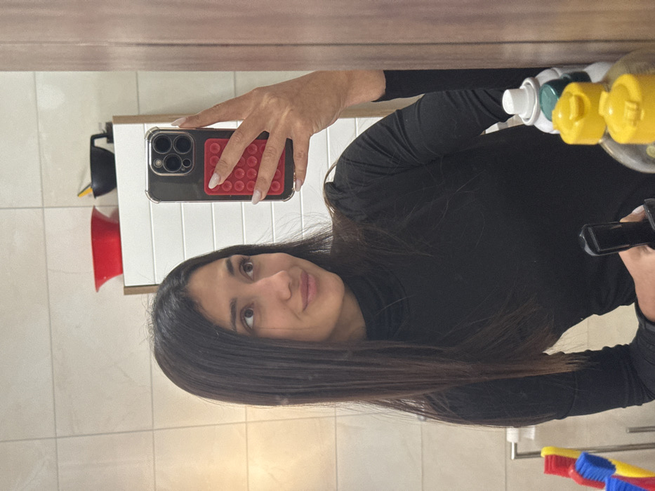 The image captures a young adult female taking a mirror selfie in an indoor setting, reportedly in the city of Cibukovac, Serbia. The subject has long, dark, straight hair and wears a black long-sleeved turtleneck shirt. She holds a dark-colored smartphone, equipped with a clear protective case and a red, multi-dimpled grip on its back, in her right hand. The phone's visible triple camera array is directed towards the mirror. The subject is looking slightly upwards and to her right, displaying a subtle smile.
The environment, visible through the mirror's reflection, consists of light-colored, square-tiled walls. Behind the subject's reflection, a white cabinet with horizontal ridging is present. On top of this cabinet, a red bowl and a black bowl containing a yellow utensil are visible. To the lower left of the reflection, a metal towel rack holds three toothbrushes: one red, one blue, and one yellow. To the lower right, several bottles, including one with a yellow cap and a white spray can with a green top, are partially visible, adjacent to a wooden cabinet or door frame. The scene is illuminated by interior lighting.