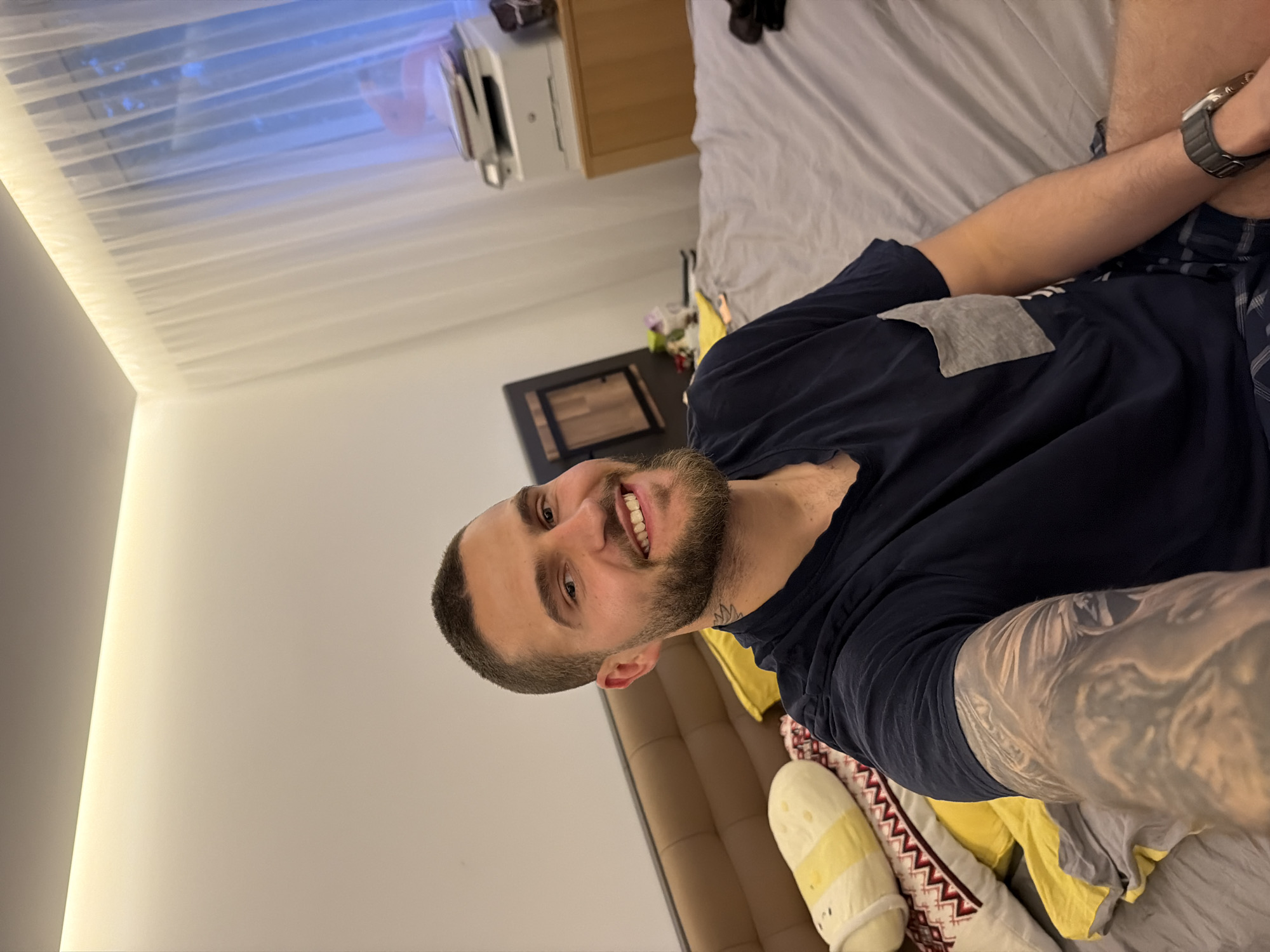 A man is lying on a bed. The man has short dark hair, a beard, and is wearing a navy blue t-shirt and plaid pants. He is also wearing a wristwatch on his left wrist, and his left arm has tattoos. The bed has a grey sheet and several pillows. The background includes a window with sheer white curtains, a desk with a printer on it, and a small black table. The location is most likely in a residential setting but is not identifiable to Sofia, Bulgaria specifically.