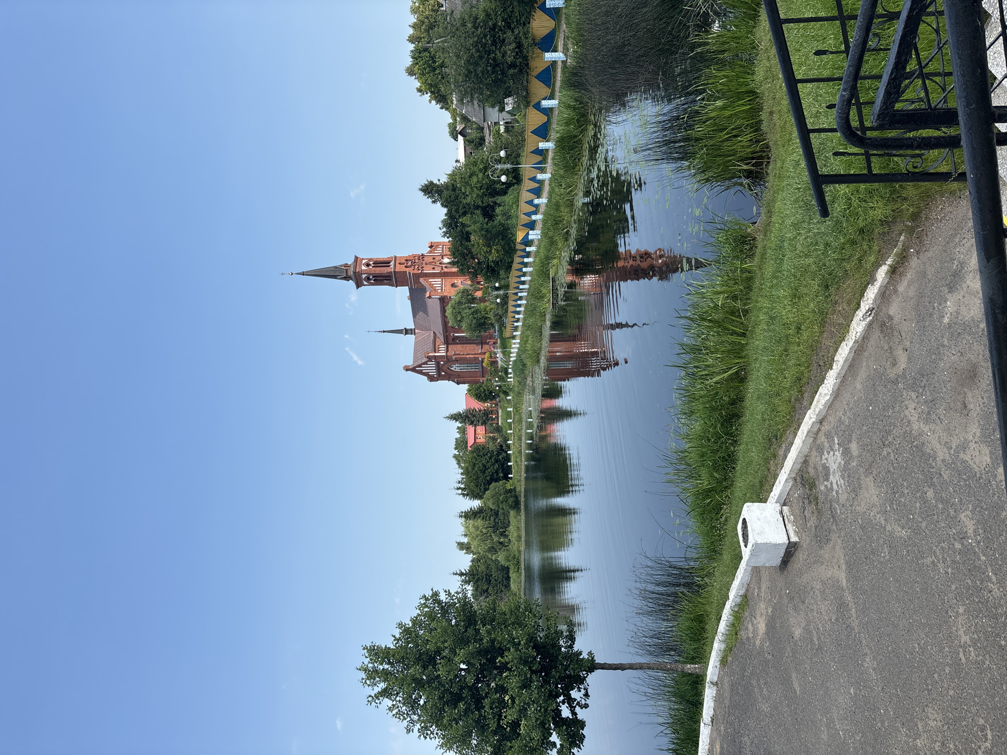 A wide-angle, eye-level shot captures a serene scene of the St. Nicholas Church in Pastavy, Belarus. The church, a grand red-brick structure with a tall, dark spire, dominates the background, its reflection shimmering in the calm, dark water of a nearby lake. The lake is bordered by lush green reeds and grass, with a narrow, yellow and blue striped fence running along its edge. A lone, leafy tree stands prominently on the left side of the frame, its branches extending over the water. The sky above is a clear, bright blue. In the foreground, a paved path curves to the right, with a white concrete planter box visible on the left. A black metal railing marks the edge of a descending staircase on the far right. The overall impression is one of peaceful, picturesque natural beauty enhanced by the presence of the historic church.