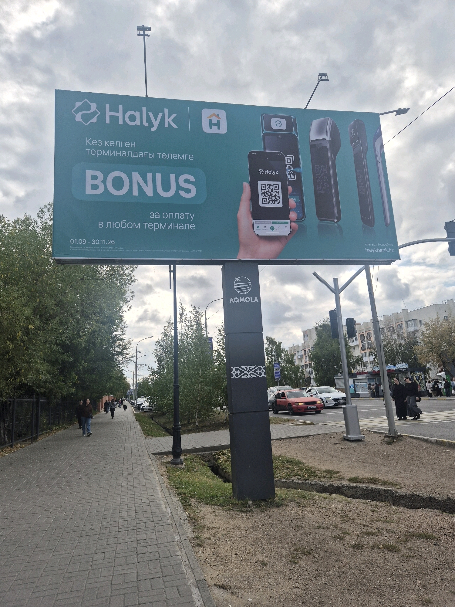 The image shows a billboard advertising a promotion by Halyk Bank in Kokshetau, Kazakhstan. The billboard features the Halyk Bank logo and text in Kazakh and Russian, promoting a bonus for payments made at any terminal. Several payment terminals and a smartphone displaying a QR code are depicted on the billboard. Below the billboard is a dark vertical sign with the "AQMOLA" logo.
The scene is a street with a pedestrian walkway on the left and a road on the right. Several people are walking on the sidewalk, and cars are driving on the road. The weather appears to be cloudy. The buildings in the background suggest an urban environment. A traffic light is visible on a pole. The presence of the "AQMOLA" logo, which is the regional transport company of Akmola region, strongly indicates the location is within Kokshetau, the administrative center of Akmola region, Kazakhstan.