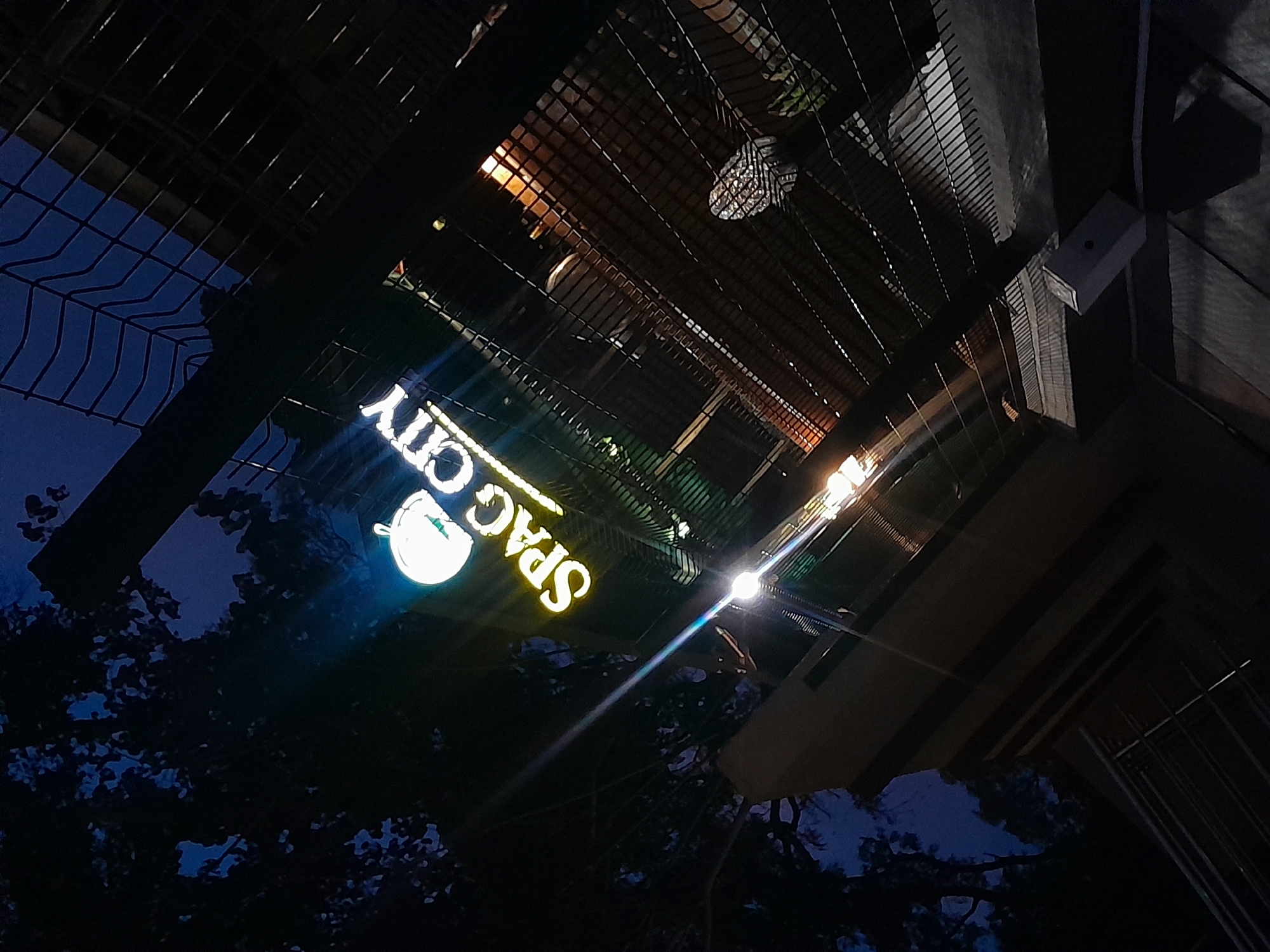 The image presents an upward-angled view of an outdoor scene at night in Ilorin, Nigeria, dominated by a brightly lit sign and an overhead metal structure. The central focus is a luminous business sign displaying the text "SPAG CITY." As viewed in the image, the text is upside down and mirrored, with "SPAG" in yellow and "CITY" in white lettering. To the left of "SPAG," a circular logo is visible, resembling a stylized bowl or a gauge, with a distinctive green element.
Above the sign and extending across the frame is an intricate metal grid or lattice structure, possibly forming a canopy or an elevated walkway. Through this structure, multiple light sources are evident. Some lights emit bright white or yellow flares, indicating exterior fixtures, while warmer, more diffused glows suggest interior lighting from a building above. The dark blue night sky is visible in the upper left, partially obscured by the silhouette of tree branches and foliage, which are also seen in the lower left against the dark background. No people or specific activities are depicted, portraying a static, commercial setting under nighttime illumination.