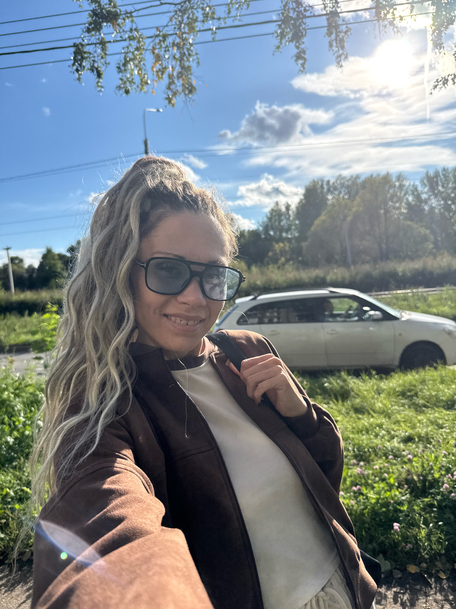 A selfie is taken outdoors in the city of Rusanovo, Russia, with a woman in the foreground. The woman has long, blonde, curly hair pulled into a high ponytail, wears dark sunglasses and a brown zip-up jacket over a light-colored top, and has braces on her teeth. She is smiling at the camera. In the background, a white car is parked on the side of a road, with trees and a grassy field surrounding it. The sky is blue with some clouds and the sun is shining brightly.