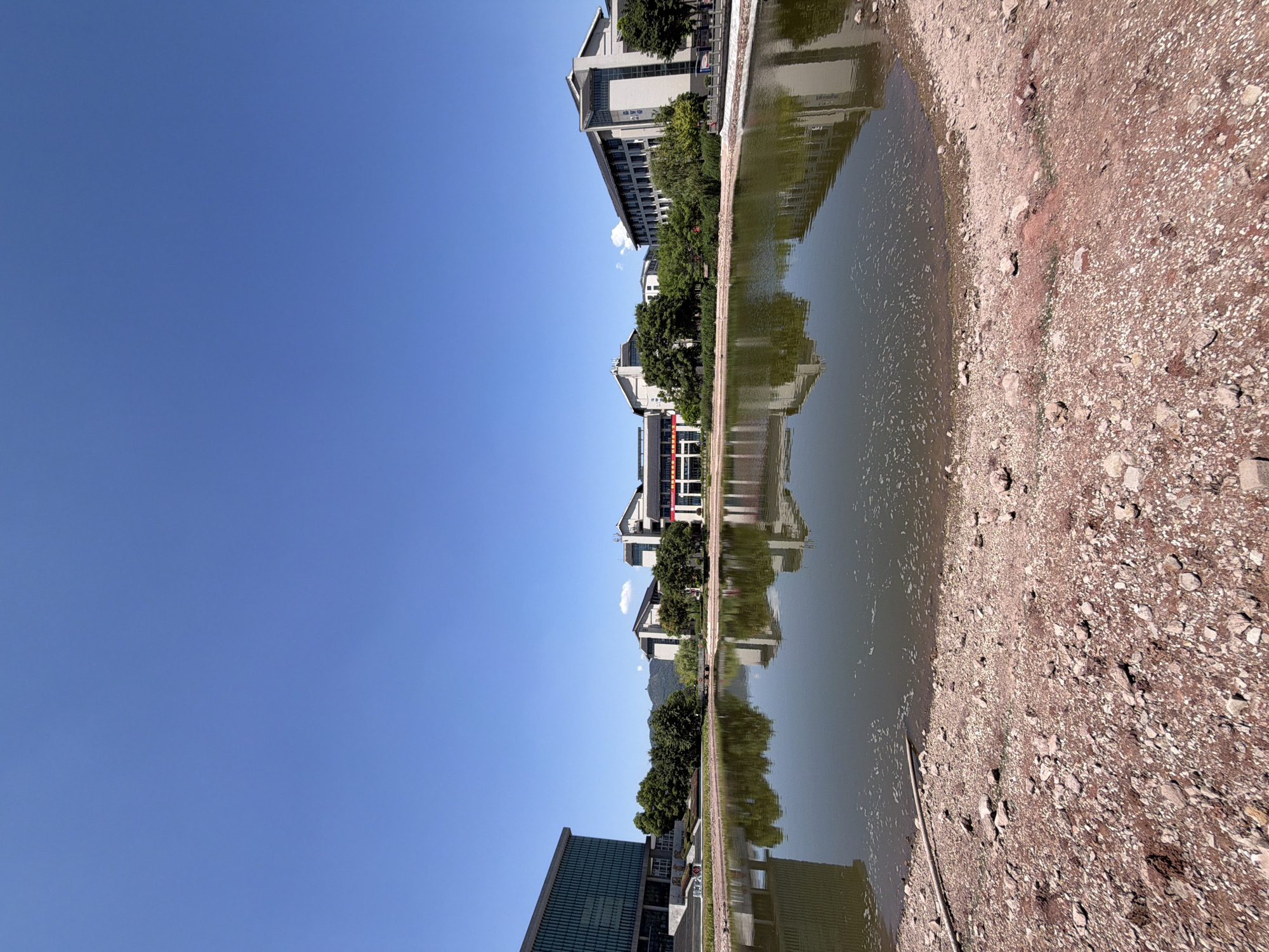The image depicts a body of water in the foreground with a rocky, dry bank. In the background, several modern buildings are visible, some with flags and banners displayed. Trees line the edge of the water and surround the buildings. The sky is clear and blue. The scene appears to be an academic campus or public area, possibly located in Jiangjin, China, given the context. The water reflects the buildings and trees.