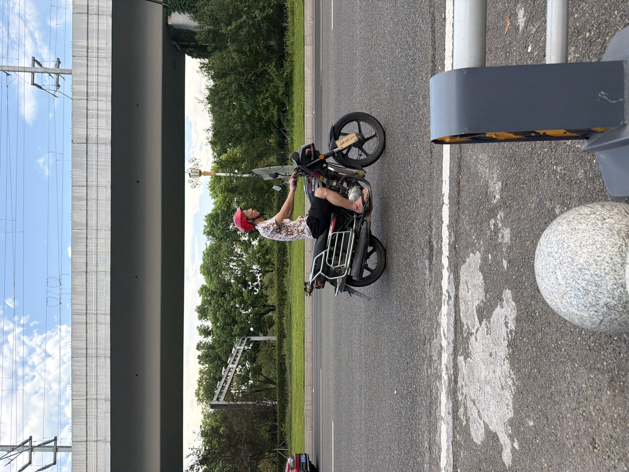 A man wearing a red helmet, a patterned short-sleeved shirt, and dark shorts rides a black motorcycle on a paved road in Jiangjin, China. The motorcycle has a windshield and a rear rack. The man is seen from his right side, and he is wearing flip-flops. In the background, a highway overpass runs horizontally across the frame. Trees and green grass are visible between the road and the overpass. The sky above is blue with scattered white clouds. A red car is partially visible on the left side of the road. A grey concrete barrier and a white line marking the road are in the foreground.
