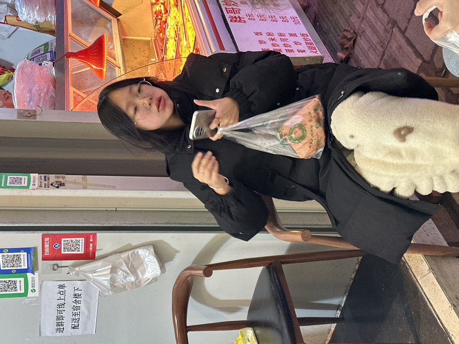 The image depicts a young East Asian female seated outside a food establishment in Jiangjin, China. She is positioned slightly right of center, wearing a black puffer jacket and round-rimmed glasses, with dark hair reaching her shoulders. Her gaze is directed towards a silver-colored smartphone held in her left hand. Her right hand grips a clear plastic bag containing a light-colored, flat food item speckled with green and red, resting on her lap. Her legs are covered by a fluffy, cream-colored garment. She sits on a wooden chair with a black seat cushion; a similar chair is partially visible to her left.
Behind the subject, on a light-colored wall or doorframe, multiple Chinese QR code payment methods (WeChat Pay) are displayed, along with a red QR code for a "red packet" promotion. A white sign with black Chinese characters hangs above a white plastic bag suspended from a hook, indicating online ordering and dormitory delivery services. To the subject's right, a glass food display case is brightly illuminated by two red heat lamps, showcasing various cooked dishes. Below the display, a white menu board with Chinese characters lists food items and prices (e.g., "饭 8元," "面 10元"). A person's head is faintly visible behind the display case in the upper right background. The bottom right corner shows a hand holding a white plastic bag. The ground is a dark surface with visible debris, including a dried leaf.