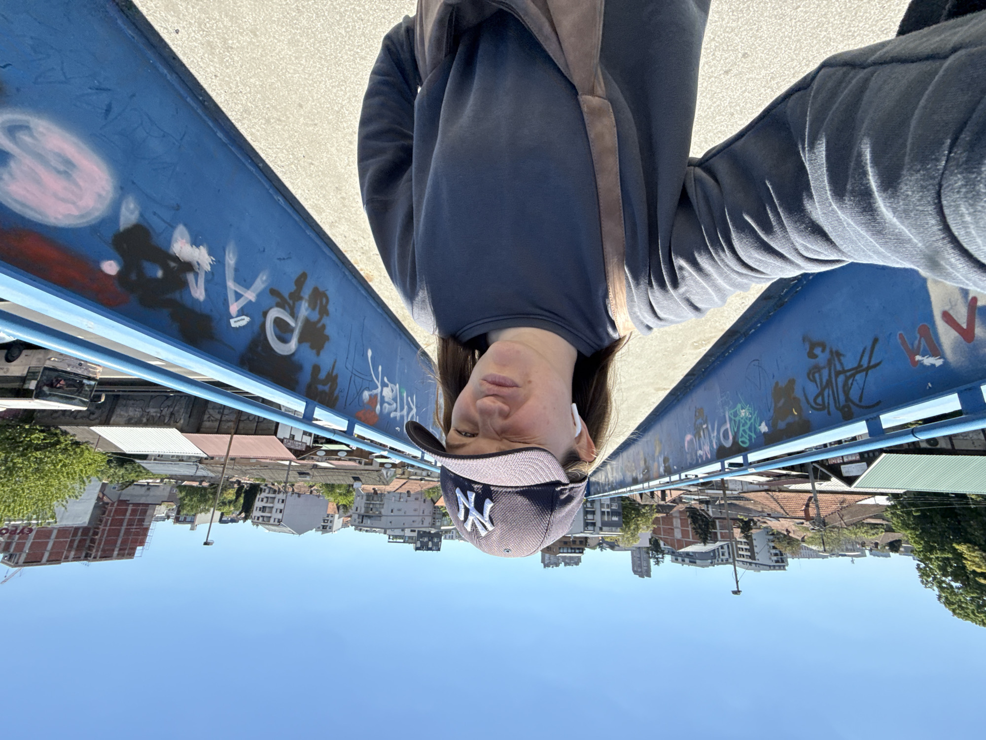 A woman wearing a dark sweatshirt, dark pants, and a New York Yankees baseball cap is on a blue pedestrian bridge. The bridge is covered in graffiti. She is facing the camera and wearing an earbud in one ear. A brown strap hangs across her chest. Below the bridge is the city of Vozdovac Urban Municipality, Serbia. Buildings and trees are visible. The sky is blue.