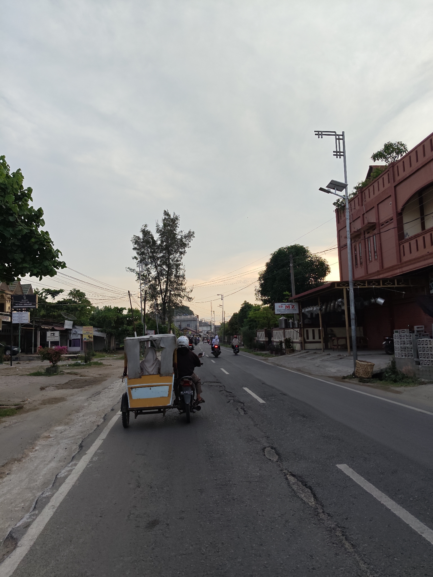 A street view in Lhokseumawe, Indonesia, shows a road stretching into the distance under a cloudy sky. A yellow motorized tricycle (becak) with two passengers is moving along the left side of the road. Several other motorcycles are further ahead. The road is paved and has white lane markings. Buildings are visible on either side of the road, including a red building on the right with a modern street light in front. Trees and vegetation line parts of the street.