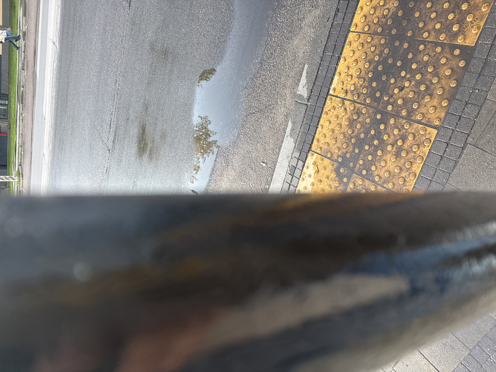 The image shows a wet asphalt road with a puddle reflecting the sky, next to yellow tactile paving for the visually impaired. The paving is bordered by gray paving stones. A blurred, dark object is in the foreground, possibly a railing or a part of a vehicle. In the distance, a building and greenery are visible, suggesting an urban environment. There is also a person visible near the green area, though they are only partially in frame.