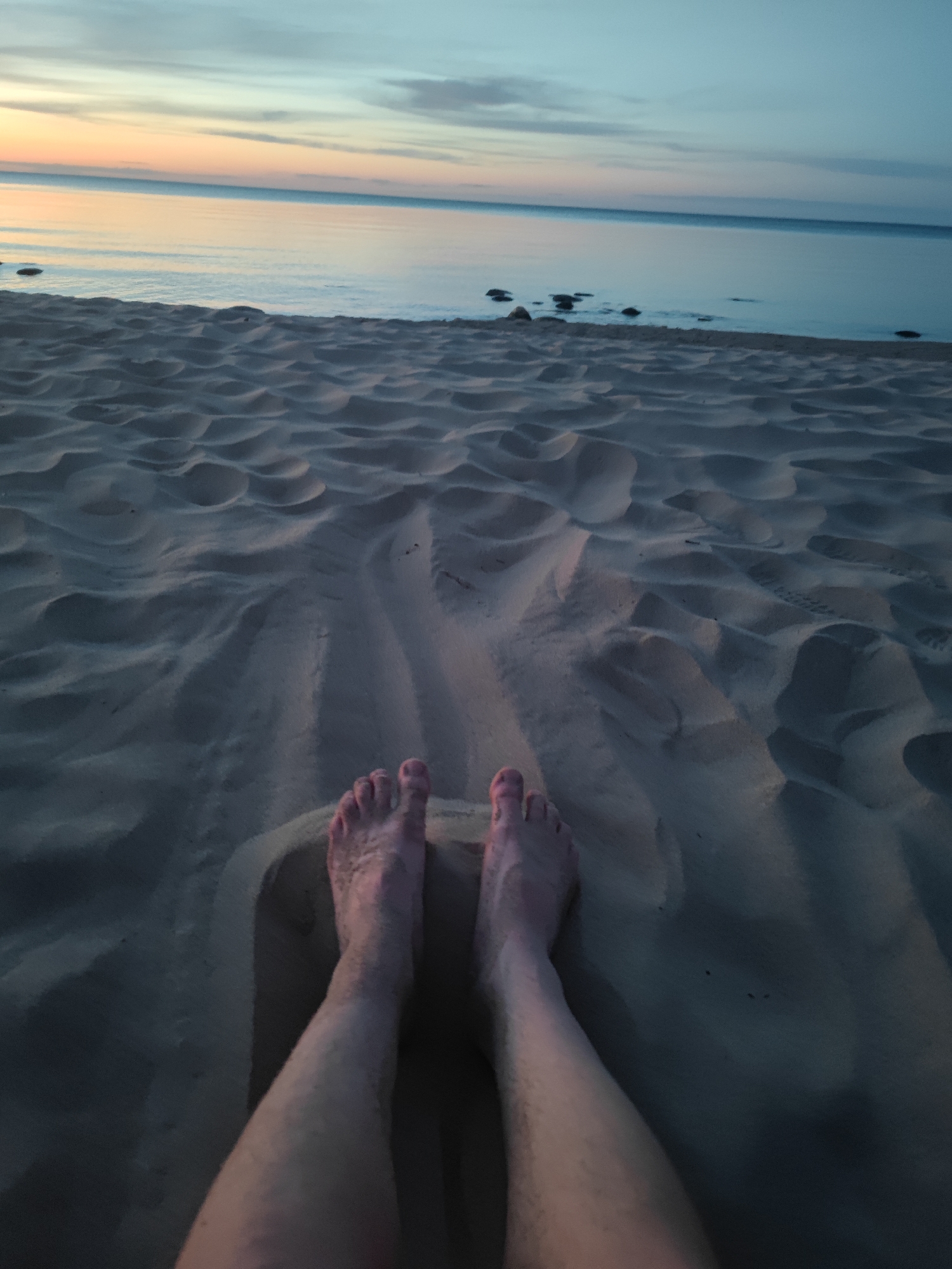 A pair of bare feet are buried in the sand on a beach. The sand has been rippled by the wind. In the background, the calm ocean meets a serene sky with hues of orange, pink, and blue, indicative of either a sunrise or sunset. Several dark rocks are visible near the waterline. The location is the beach of Motornoe, Russia.