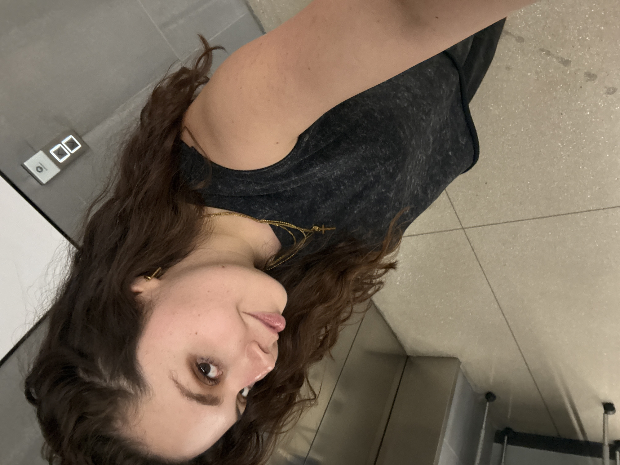 A young woman is captured in a selfie-style photograph, shown from the chest up, with her head tilted slightly and eyes directed towards the camera. She has long, wavy dark brown hair flowing over her shoulders and wears a dark grey or black marled sleeveless top. A delicate gold chain necklace, featuring a small cross-like pendant, is visible around her neck, and a small gold earring adorns her left ear. Her expression is calm and subtle, with a hint of a smile or thoughtful gaze, and her lips are slightly parted.
The setting appears to be an indoor space, likely a hallway or elevator lobby within a modern building. The background features a light-colored, possibly speckled terrazzo or tiled floor to her right, and a dark grey wall behind and to her left. A prominent digital display panel is mounted on the wall, showing the numbers "00" along with a downward arrow and a small circular icon, suggesting an elevator floor indicator. The overall lighting is consistent with indoor artificial illumination. The image context places the location in Mexico City, Mexico.