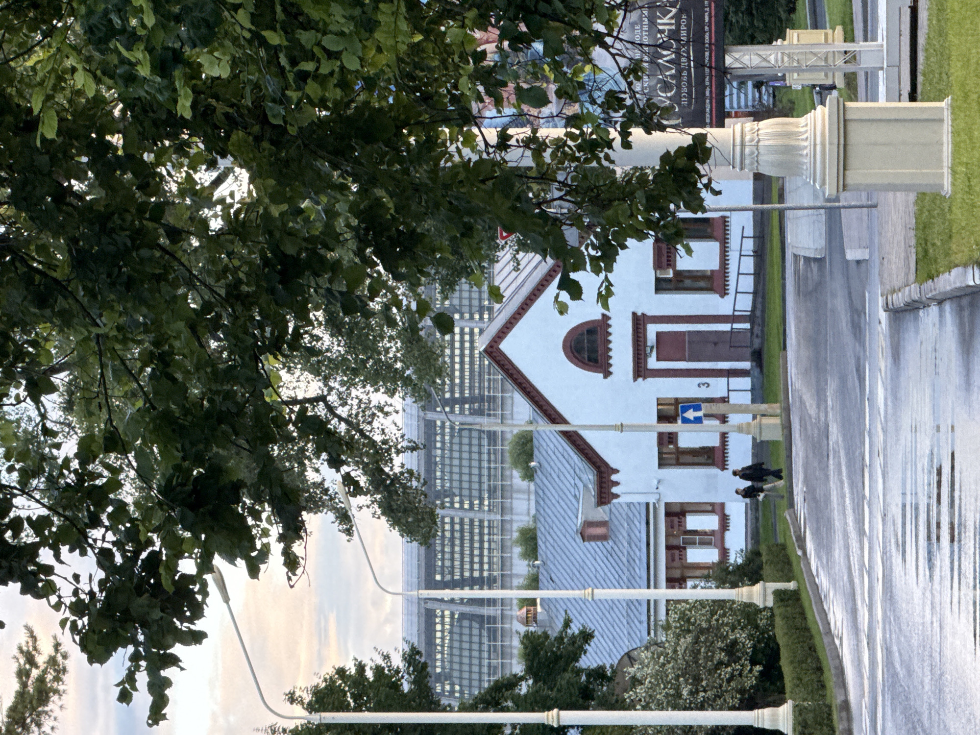 A street view shows a crosswalk on a wet asphalt road in front of a white building with a red-trimmed roof. Two pedestrians walk across the crosswalk. Trees with green leaves overhang the scene, partially obscuring a modern glass building in the background. A lamppost with decorative detailing stands on the right side of the road. The location appears to be Moscow, Russia, with a sign in Cyrillic visible in the background, potentially advertising an event or business.