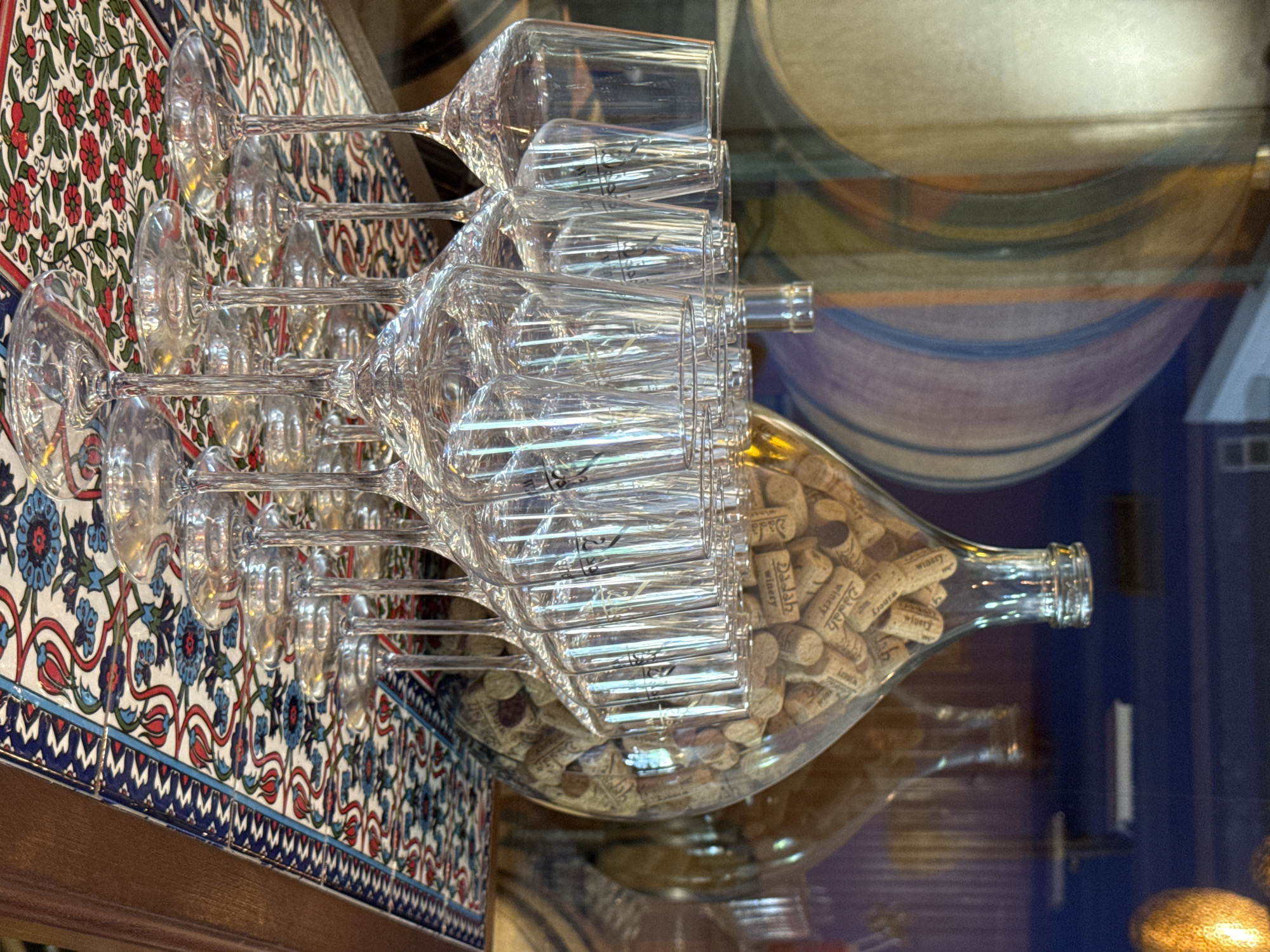 In Hof HaCarmel Regional Council, Israel, a close-up shot focuses on a collection of clear wine glasses arranged on a decorative tiled surface. The tiles feature a vibrant pattern of red flowers, blue floral designs, and white swirls with green accents. Behind the glasses, a large, transparent glass bottle is filled with numerous wine corks, some of which bear the imprint "Dalah Winery". To the left and right of this bottle, other glass bottles are visible, though less clearly defined. In the background, a portion of what appears to be a wine barrel with painted bands of color is reflected in the glass. The overall scene suggests a winery or tasting room setting.