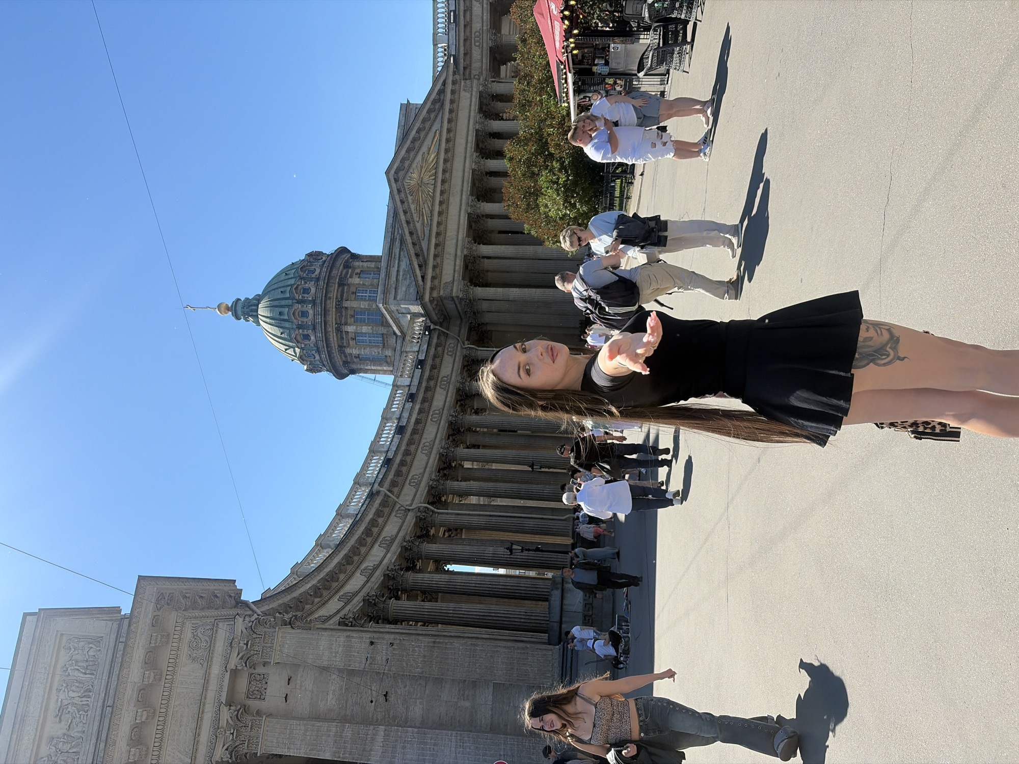 The image captures a sunny day in front of the Kazan Cathedral in Saint Petersburg, Russia. A young woman with long, dark hair, wearing a black crop top and a black pleated skirt, is in the foreground, extending her right hand towards the camera. Her left hand is holding a leopard-print bag. She has a tattoo visible on her left thigh.
In the background, the grand facade of the Kazan Cathedral, with its semicircular colonnade and prominent green dome topped with a cross, is visible under a clear blue sky. Several people are walking around the square, including a woman in a leopard-print top and jeans to the left of the main subject, and a couple standing closer to the cathedral on the right. A food stall with a red awning is also present on the right side of the frame. The ground is paved, and shadows are cast by the people and structures.