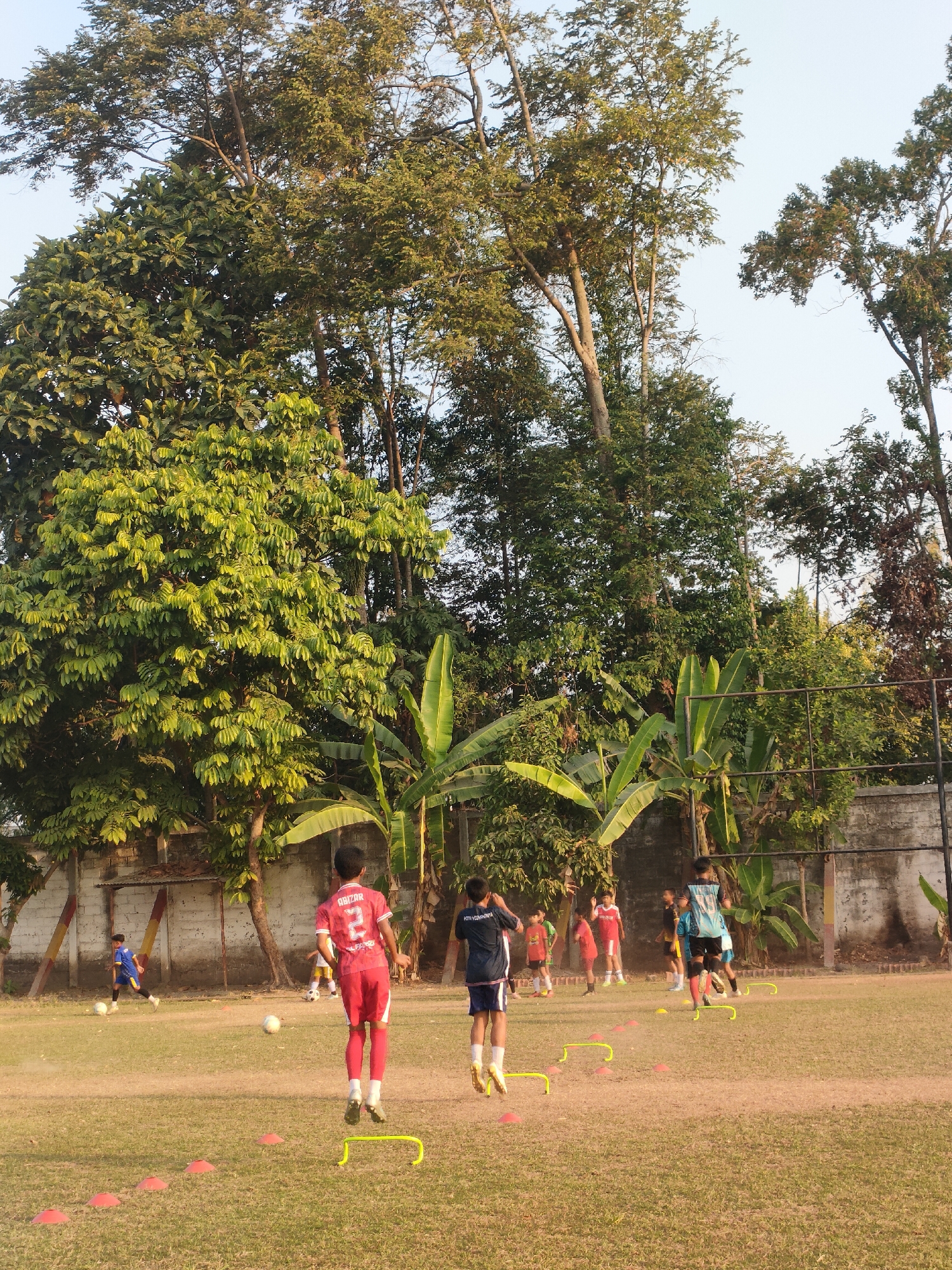 In Banguntapan, Indonesia, a group of young boys are practicing soccer on a grassy field. The field is marked with orange cones and yellow agility hurdles, indicating a training session. Several boys are in motion, dribbling, running, or jumping over obstacles. One boy in a red jersey with the number 2 is in the foreground, mid-jump over a yellow hurdle. Another boy in a blue and white jersey is also jumping over a hurdle. In the background, other boys are scattered across the field, some with soccer balls. The field is surrounded by lush green trees and foliage, with a brick wall in the background. The sky above is a clear, pale blue.
