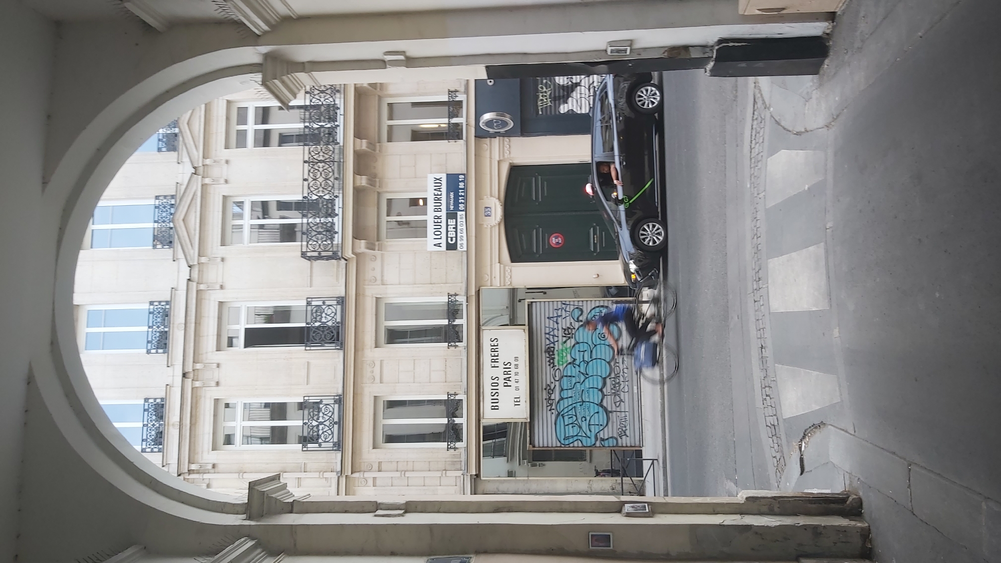 The image captures a street view in Paris, France, as seen through a large archway. The building facade across the street features multiple stories with windows, some with decorative balconies. A sign reading "A LOUER BUREAUX" (Offices for Rent) is visible on the upper floor. Below this, another sign indicates "BUSIOS FRERES PARIS" with a telephone number. The ground floor shows a closed shutter with graffiti. In front of the building, a person is cycling past, their motion blurred. To the right, a black car is parked or moving slowly, with its passenger door open and a person leaning out. The street includes a pedestrian crossing. The overall impression is of a typical Parisian street scene with commercial activity and urban life.