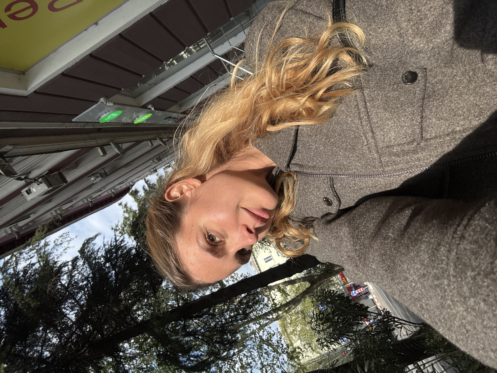 An outdoor portrait-style image features a woman with long, wavy blonde hair and fair skin. She is looking slightly upwards towards the camera with a neutral to subtly pleasant expression. She is wearing a grey, textured jacket or coat with a visible black zipper and a dark button on a small pocket flap.
The background suggests an urban environment. Above the woman, there's a building's underside or awning, detailed with white trim and brown paneling. Two circular green illuminated indicators or buttons are mounted on a grey panel beneath this structure. A yellow sign with partially visible red text, including the letters "e" and "o," is also present, possibly indicating a business.
Below and behind the woman, numerous green trees with dense foliage are prominent, suggesting a park or street-lined area. Through the trees, patches of clear blue sky with some white clouds are visible. Further in the background, a street can be seen with several vehicles, including a red truck, and various street signs, though their specific details are indistinct. The strong natural light and pronounced shadows indicate it is daytime on a sunny day.