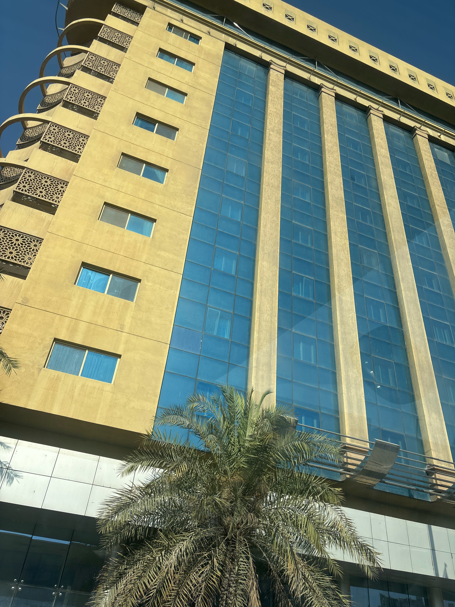The image depicts a low-angle view of a modern, multi-story building with a yellow facade and large blue glass windows. The building has a distinctive architectural style with patterned screens on the balconies on the left side, and vertical concrete elements separating the glass sections on the right. A palm tree is visible in the foreground, its fronds partially obscuring the lower portion of the building, which features a reflective glass exterior. The sky is clear and blue. The building appears to be located in the city of Dhahran, Saudi Arabia, a region known for its contemporary architecture and presence of palm trees. There are no people or animals visible in the image. The scene is static and showcases the building's facade against a clear sky.