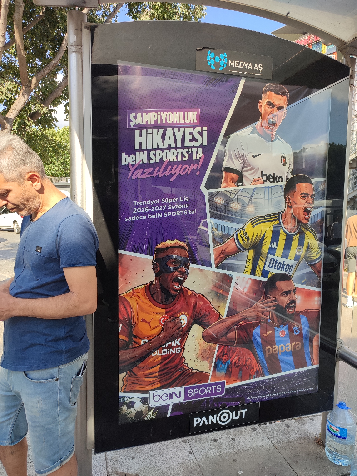 An outdoor advertising display for beIN SPORTS is seen at a bus stop in Kadıköy, Turkey. The display features a collage of illustrated football players in action. A man in a blue t-shirt and denim shorts stands to the left of the advertisement, looking down at his phone. The advertisement itself is vibrant and predominantly purple and white, with text in Turkish that translates to "Championship Story is written on beIN SPORTS!". Beneath this headline, smaller text specifies "Trendyol Süper Lig 2026-2027 Season, only on beIN SPORTS!". The bottom of the advertisement includes the "PANOUT" logo and a disclaimer stating "Images and visuals created using artificial intelligence technology." A large bottle of water is placed on the ground to the right of the display. The bus stop structure has a metallic grey finish, and the surrounding environment suggests an urban setting with trees and a blue sky visible.